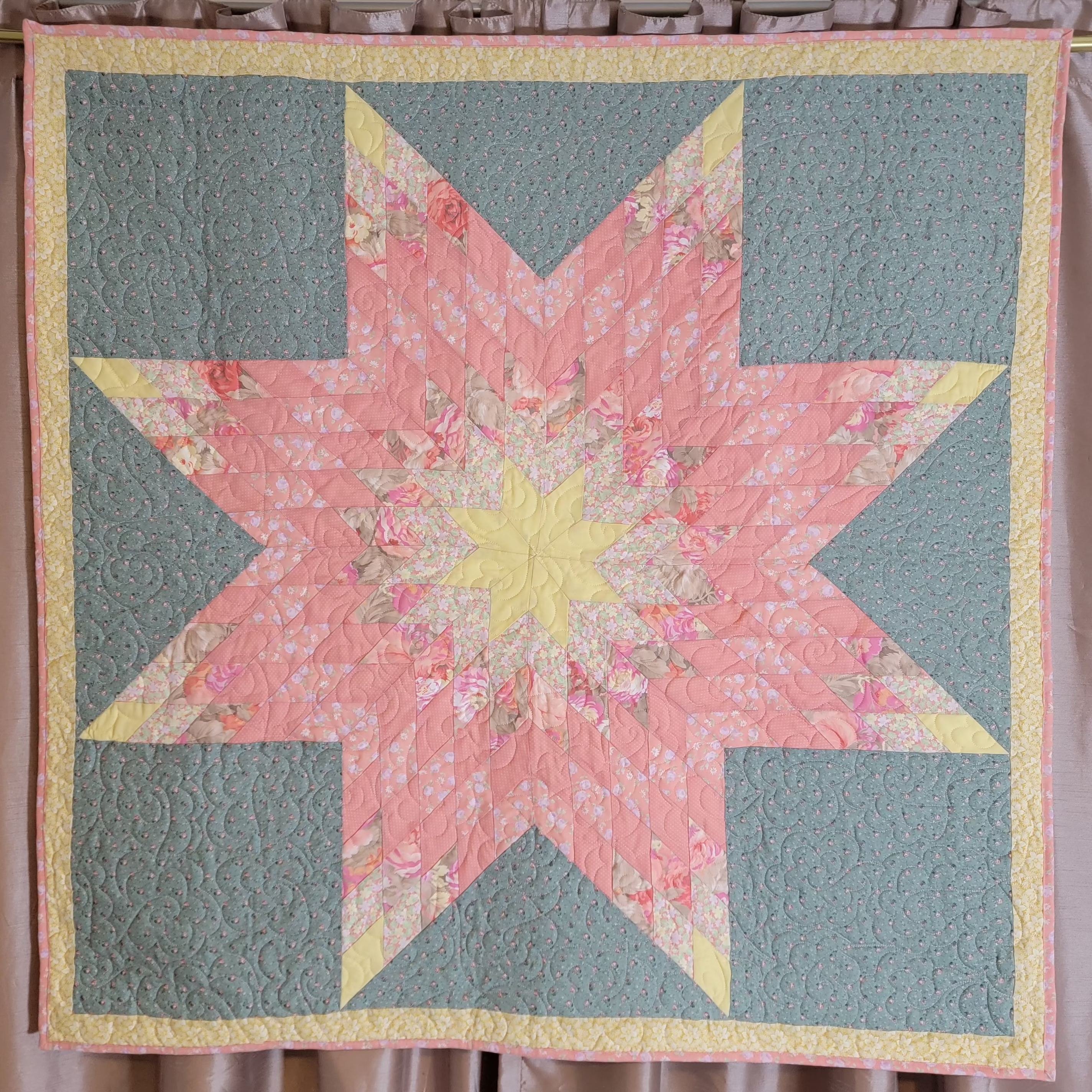 A handmade quilted starburst quilt hanging on a rod against a curtain backdrop. The quilt features a central pink floral star with a yellow center, surrounded by yellow triangles and four muted sage green corners.