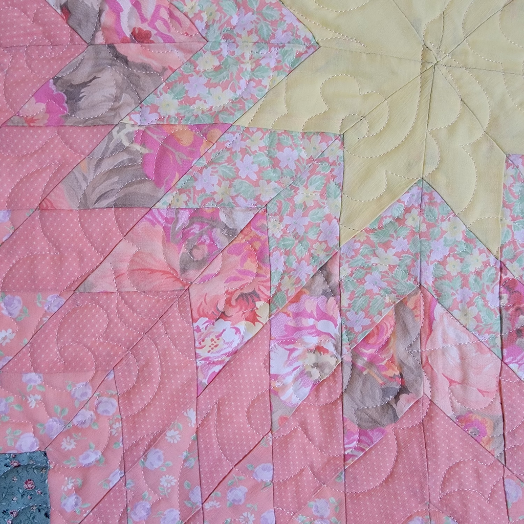 A close-up of a quilted fabric featuring a geometric starburst pattern in shades of pink, floral prints, and a pale yellow section.