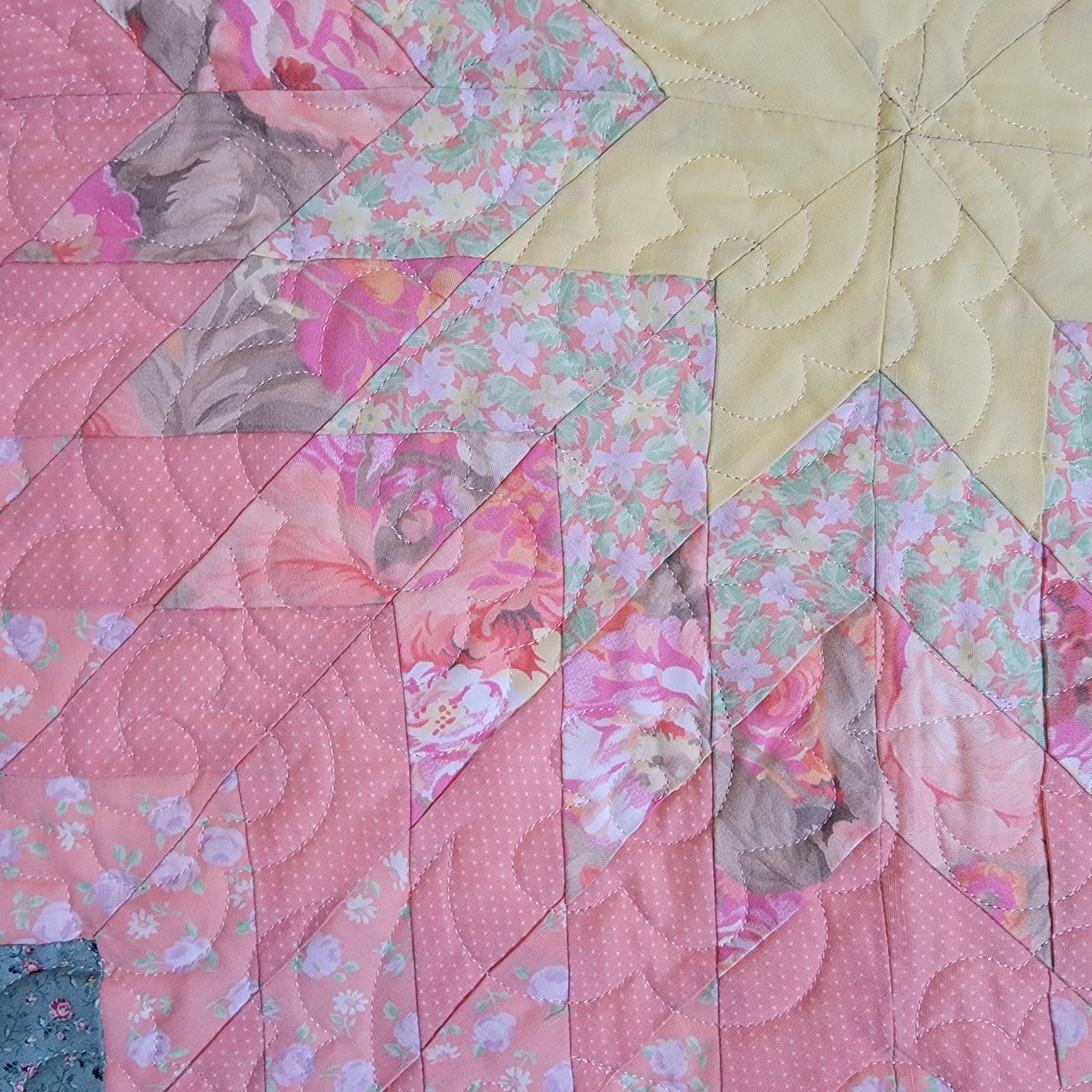 A close-up of a quilted fabric featuring a geometric starburst pattern in shades of pink, floral prints, and a pale yellow section.