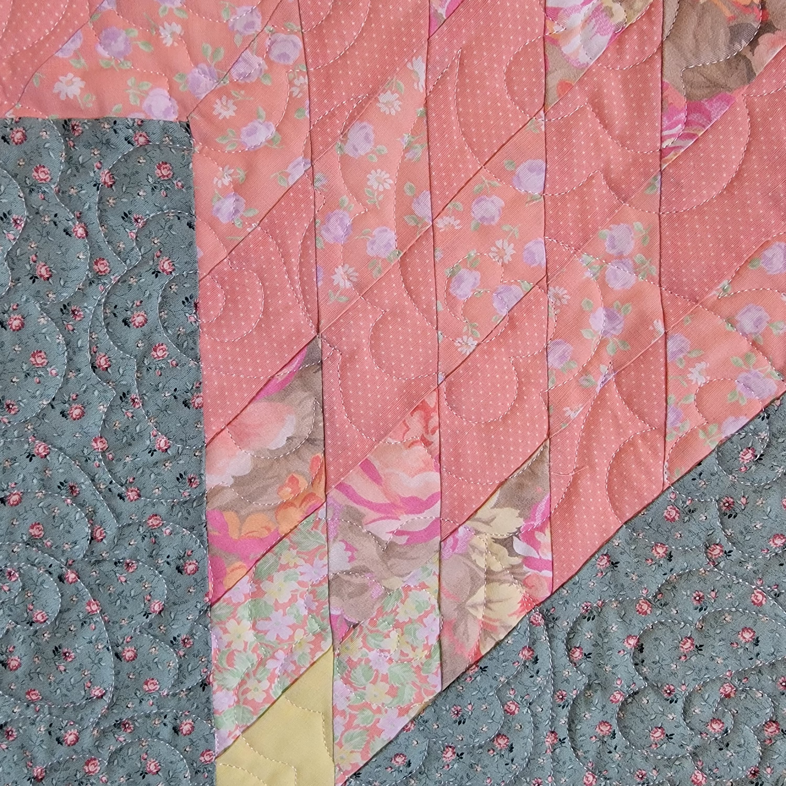 A close-up of a quilted fabric panel with diagonal stitched sections in pink floral print, dusty blue with small pink floral motifs, pale yellow, and a light beige border.
