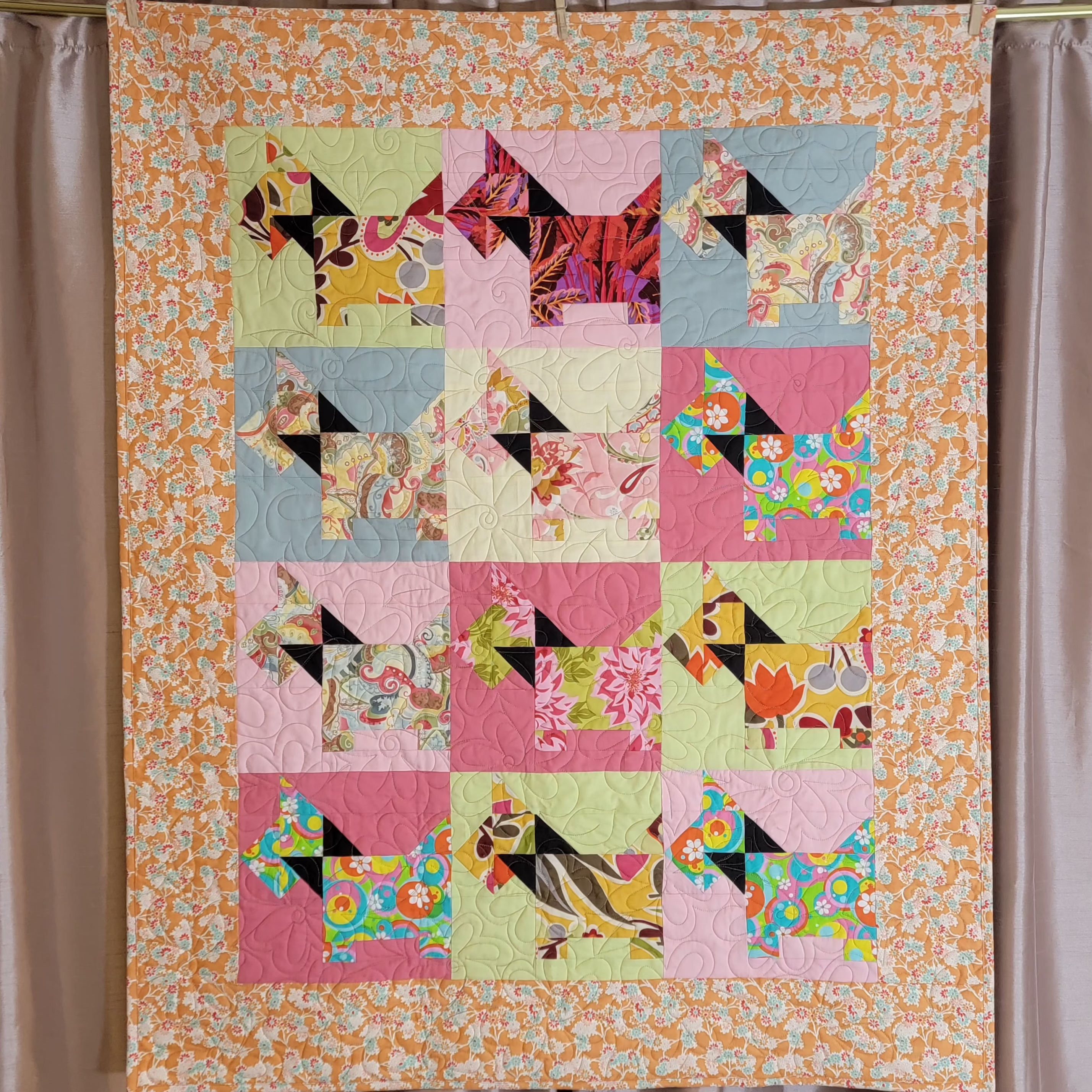 A handmade quilt featuring a grid of colorful dog-shaped patches with floral and geometric patterns, bordered by an orange and white floral print. The quilt is hung on a curtain rod with wooden clips against a brown curtain backdrop.