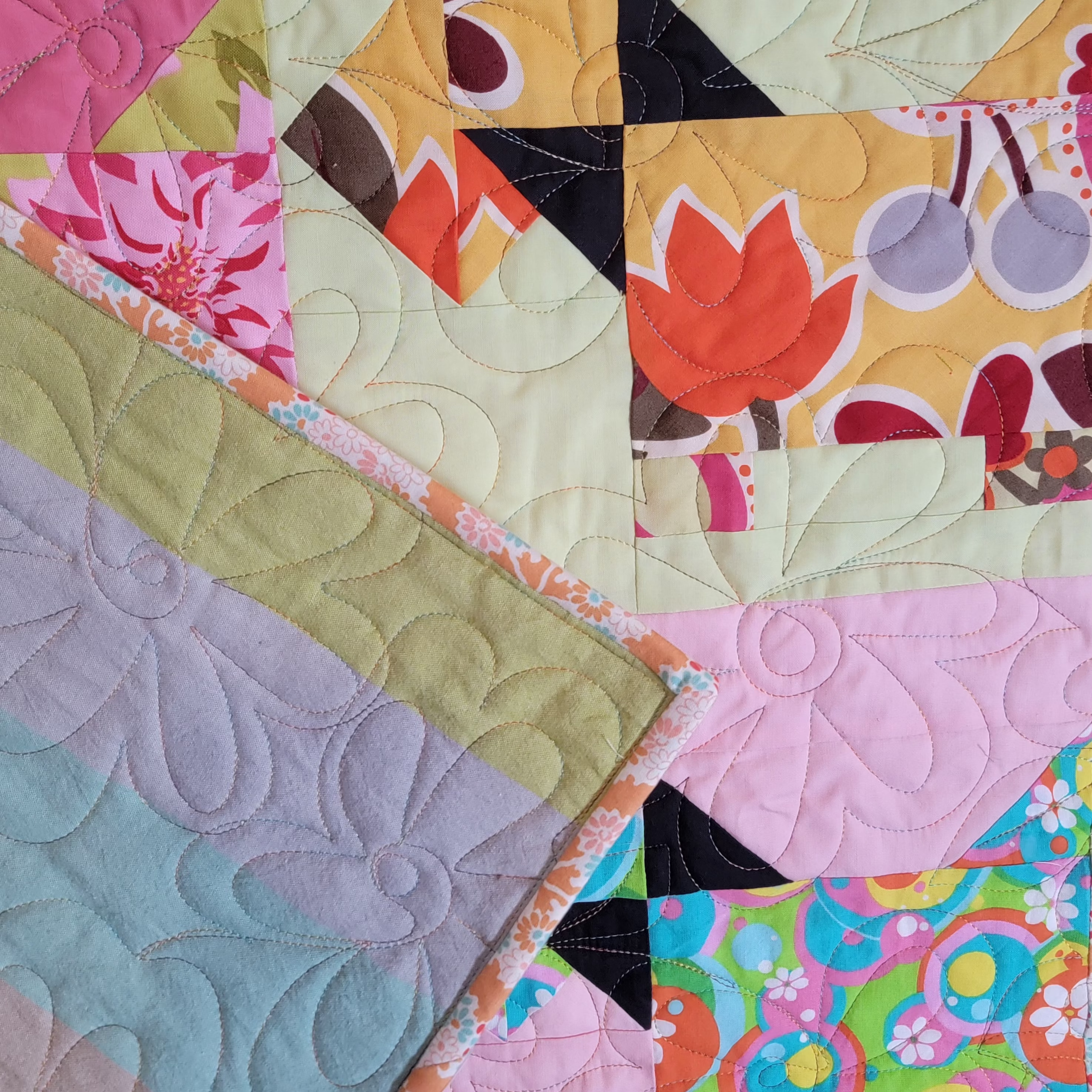 A close-up of a colorful quilted fabric with multiple patterned and solid fabric blocks, including floral prints, geometric shapes, and swirling stitched textures.