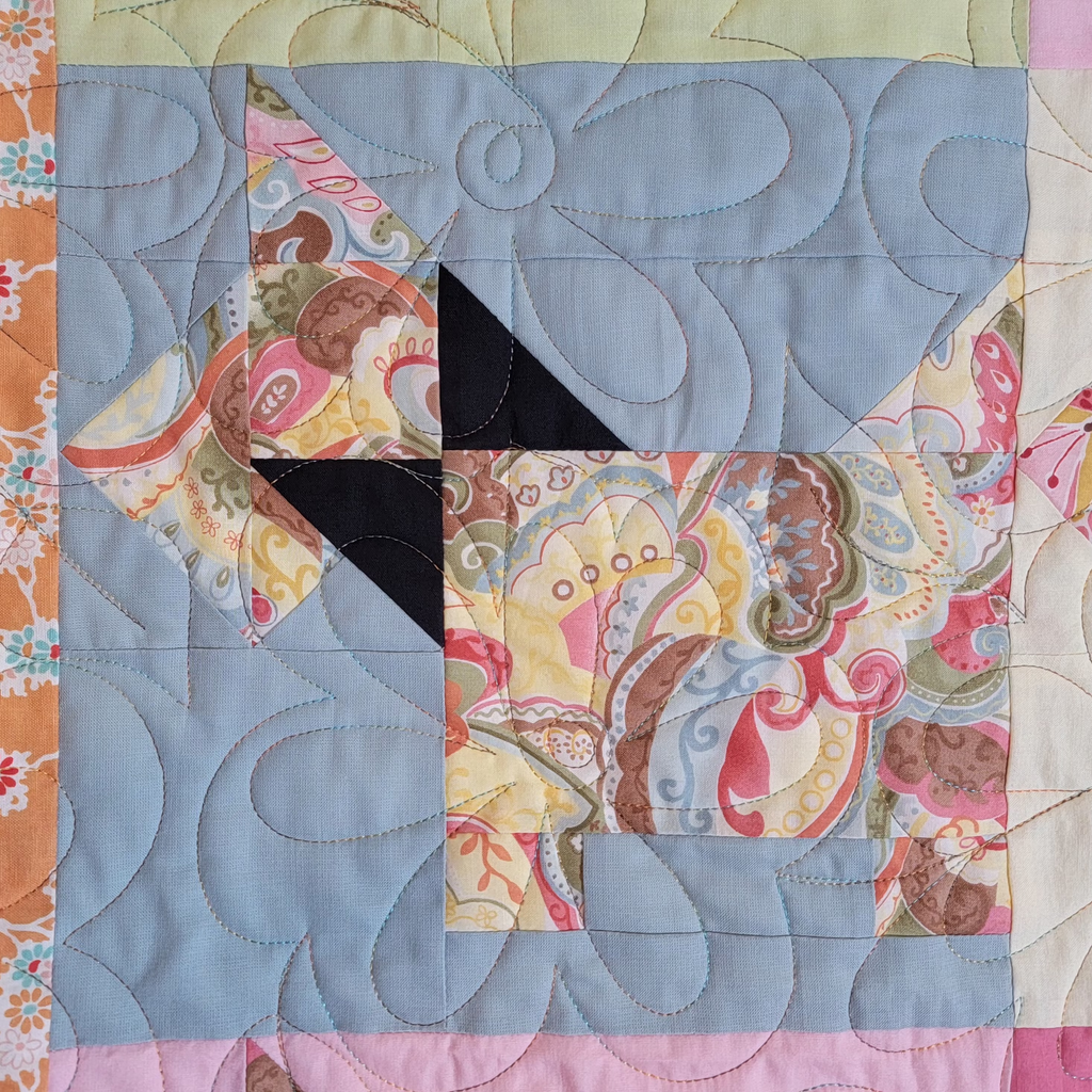 A close-up of a quilted fabric block featuring a stylized dog made from patterned fabrics in pink, beige, yellow, and brown, with a light blue background and swirling stitched quilting.