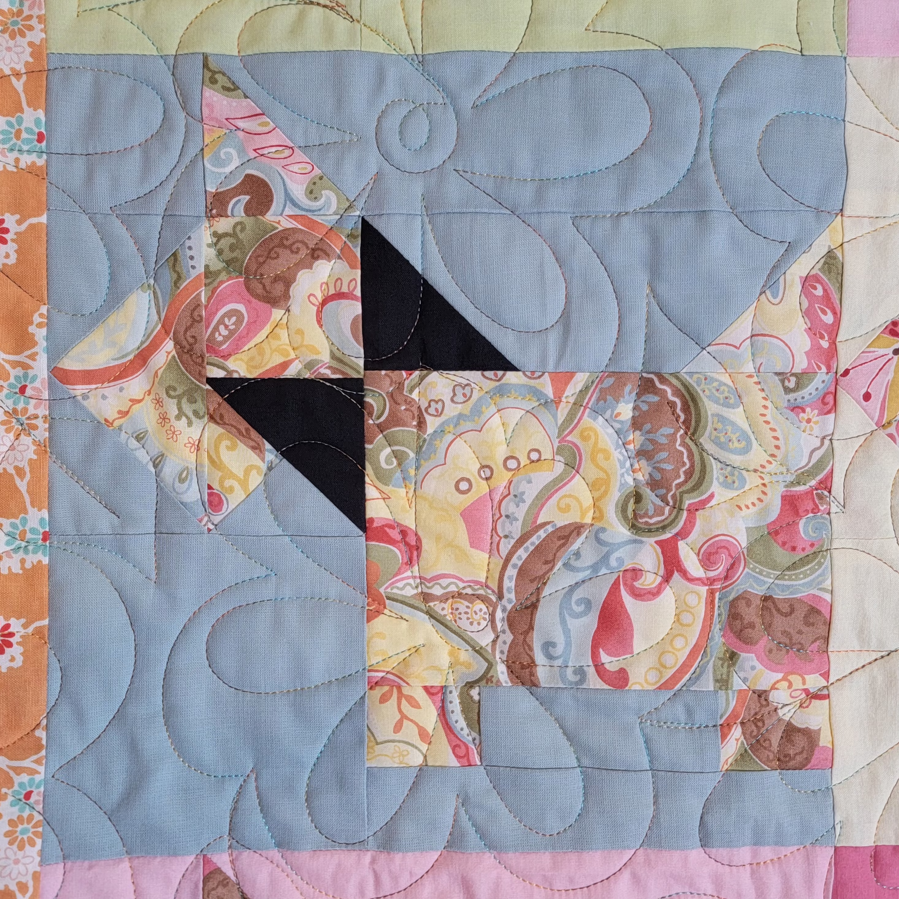 A close-up of a quilted fabric block featuring a stylized dog made from patterned fabrics in pink, beige, yellow, and brown, with a light blue background and swirling stitched quilting.