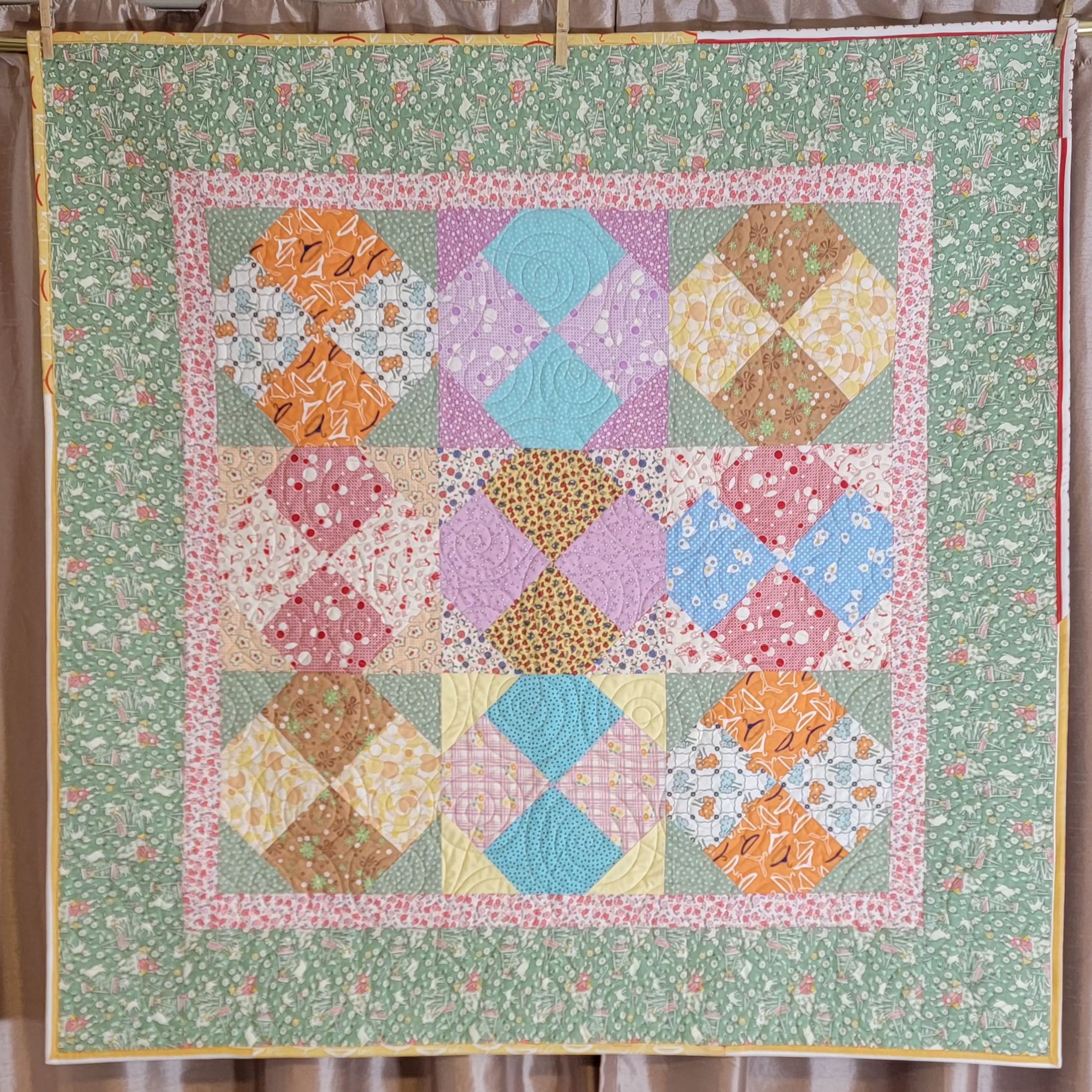 Quilt hung up on a rod, the quilt is made up of pastel green, pink and blue tones in multiple prints and patterns. 