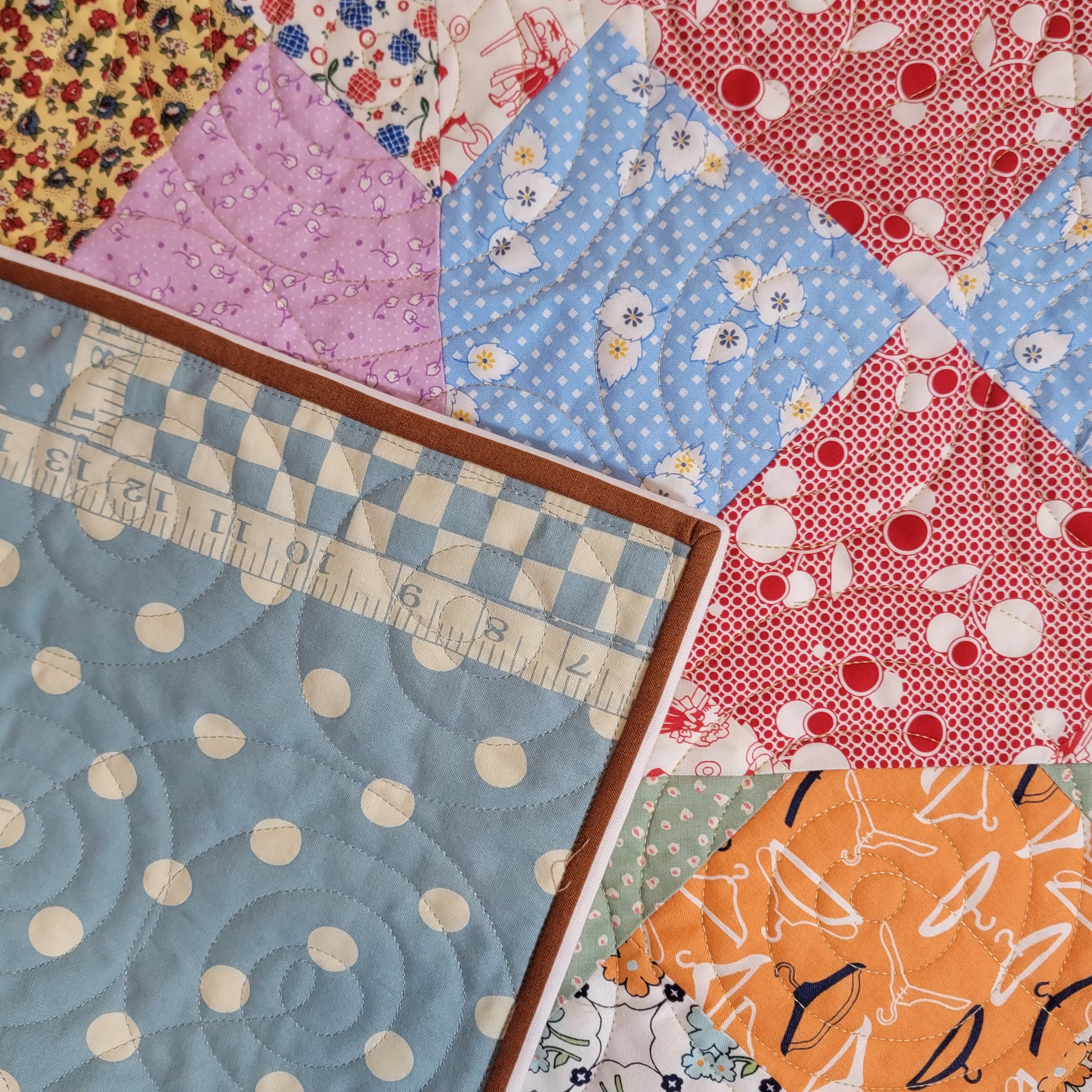 Close up of a handmade quilt. The main side is made up of pastel blues, orange, pinks and reds in multiple prints and patterns. The reverse side is showing which is a polkadot print with a ruler print along the edge in a baby blue colour with a brown trim edge. 