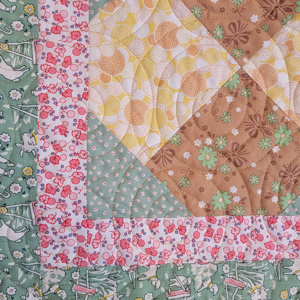 A close up of a handmade quilt with a diamond shape made up of pastel brown and yellow prints, bordered with a pink and green pastel print. 