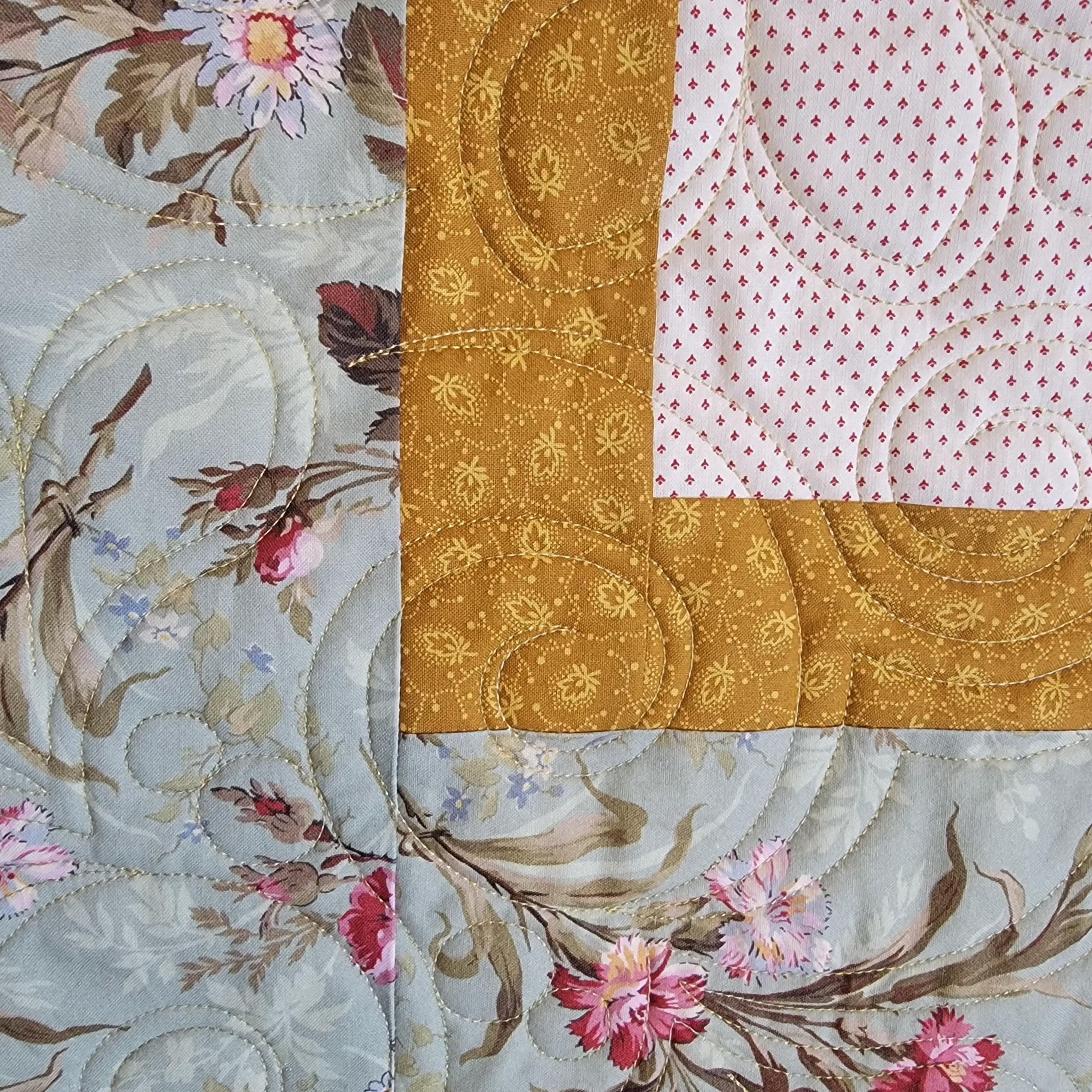 A close-up of a quilted textile showing three fabric sections: a pale blue floral print with pink flowers and brown leaves, a wide mustard-gold border with floral embroidery, and a white section with small pink dots.