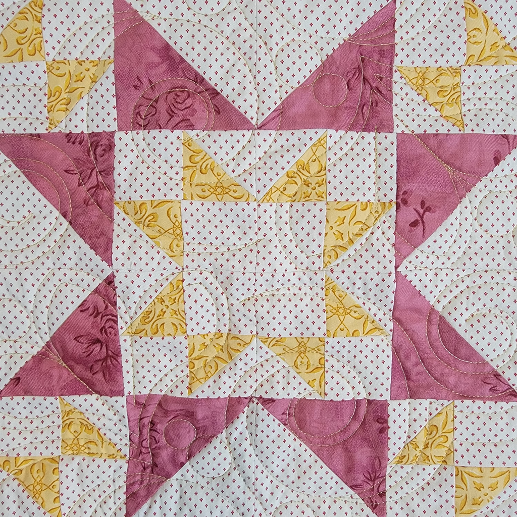 A close-up of a quilted fabric featuring a geometric star pattern made of white fabrics with tiny pink dots, dusty pink triangles, and mustard yellow triangles.