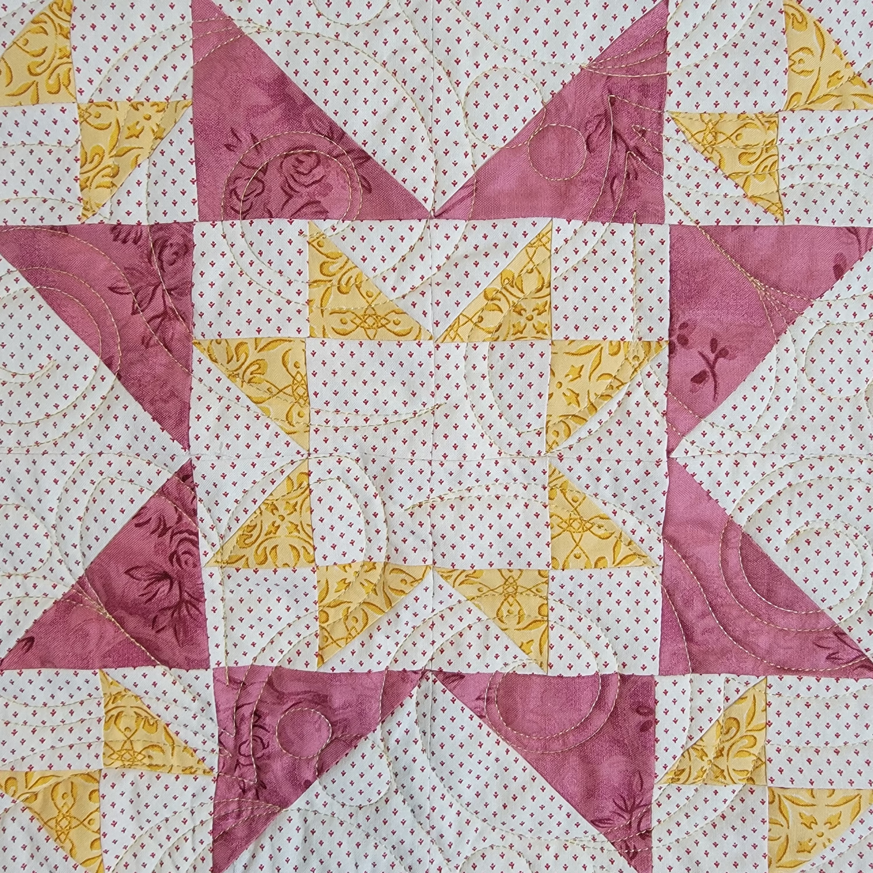 A close-up of a quilted fabric featuring a geometric star pattern made of white fabrics with tiny pink dots, dusty pink triangles, and mustard yellow triangles.
