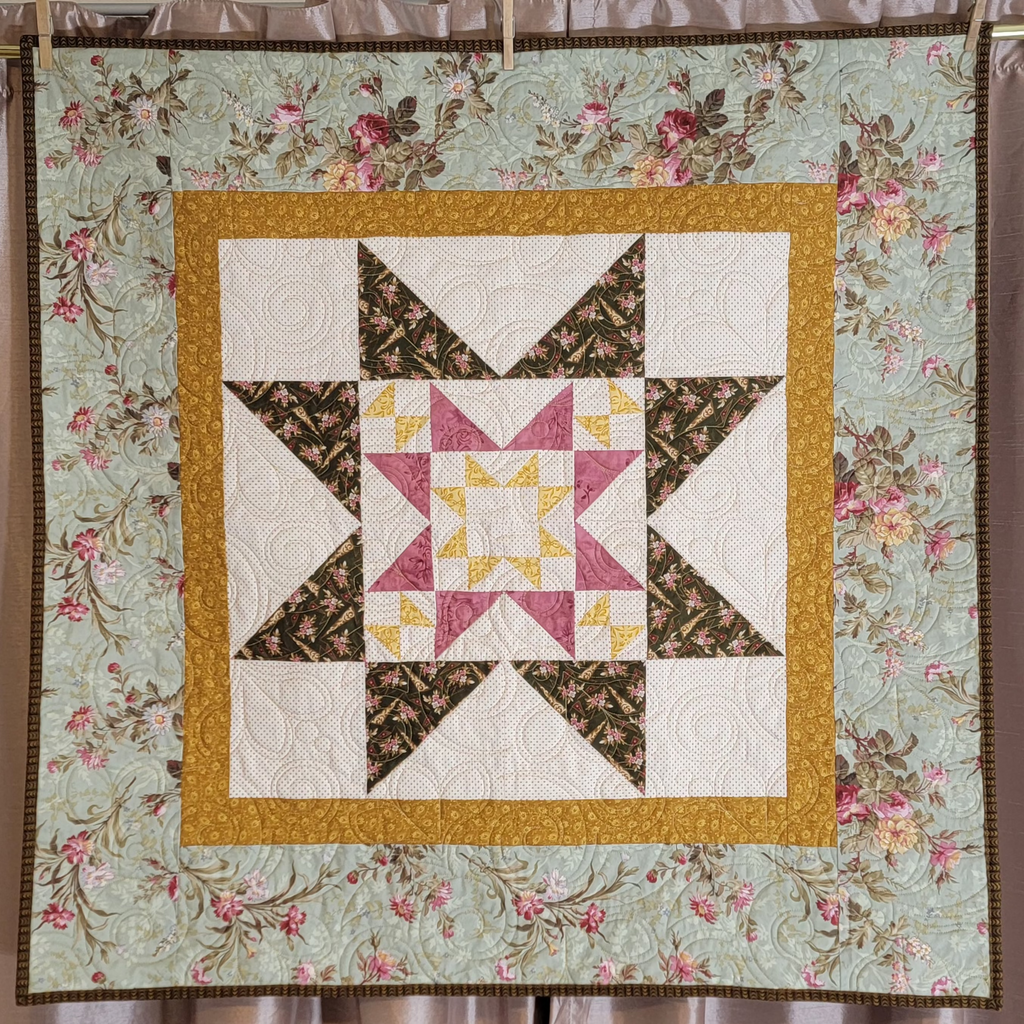 A square quilt with a floral border and a central starburst patchwork design, displayed hanging from clips against a curtain backdrop.