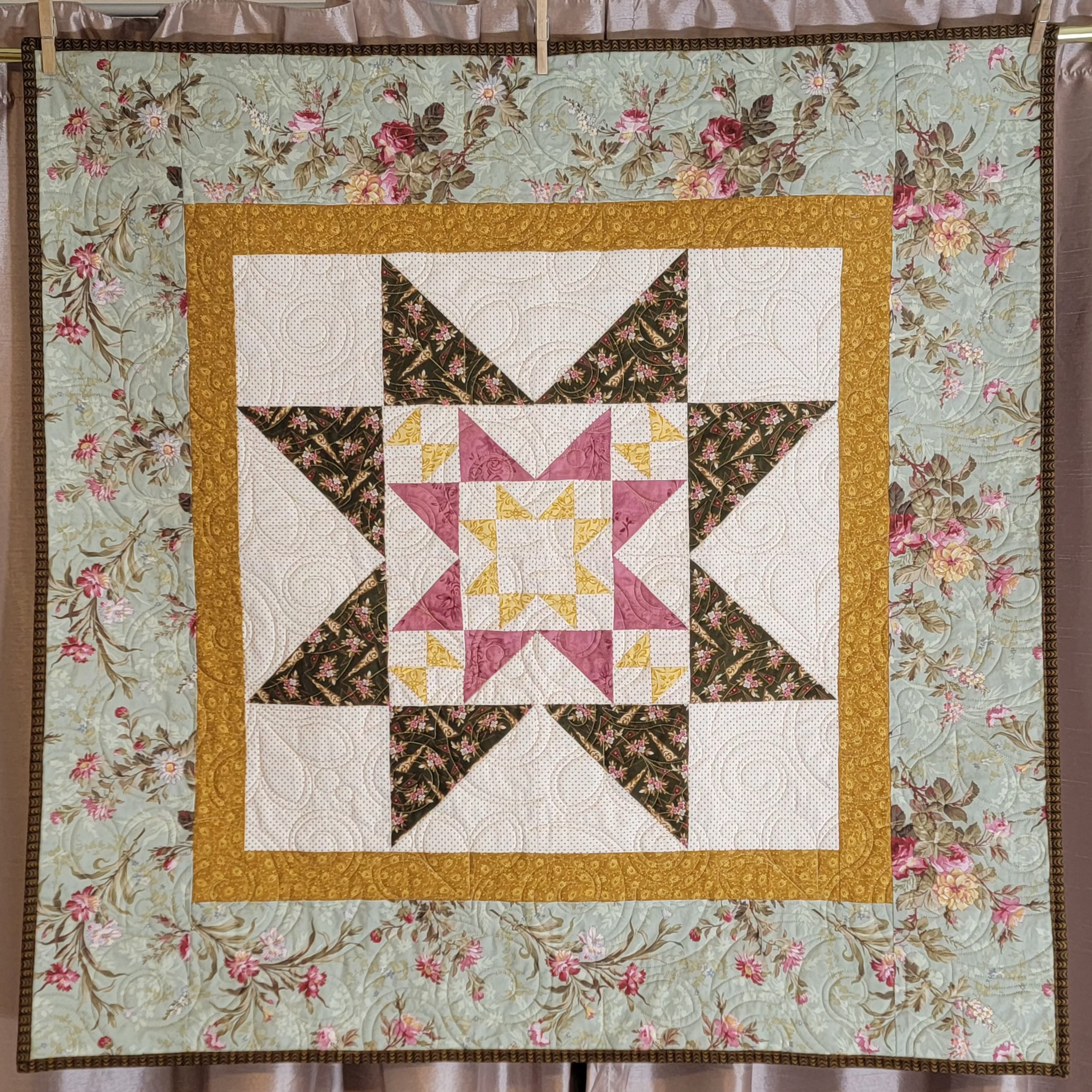 A square quilt with a floral border and a central starburst patchwork design, displayed hanging from clips against a curtain backdrop.