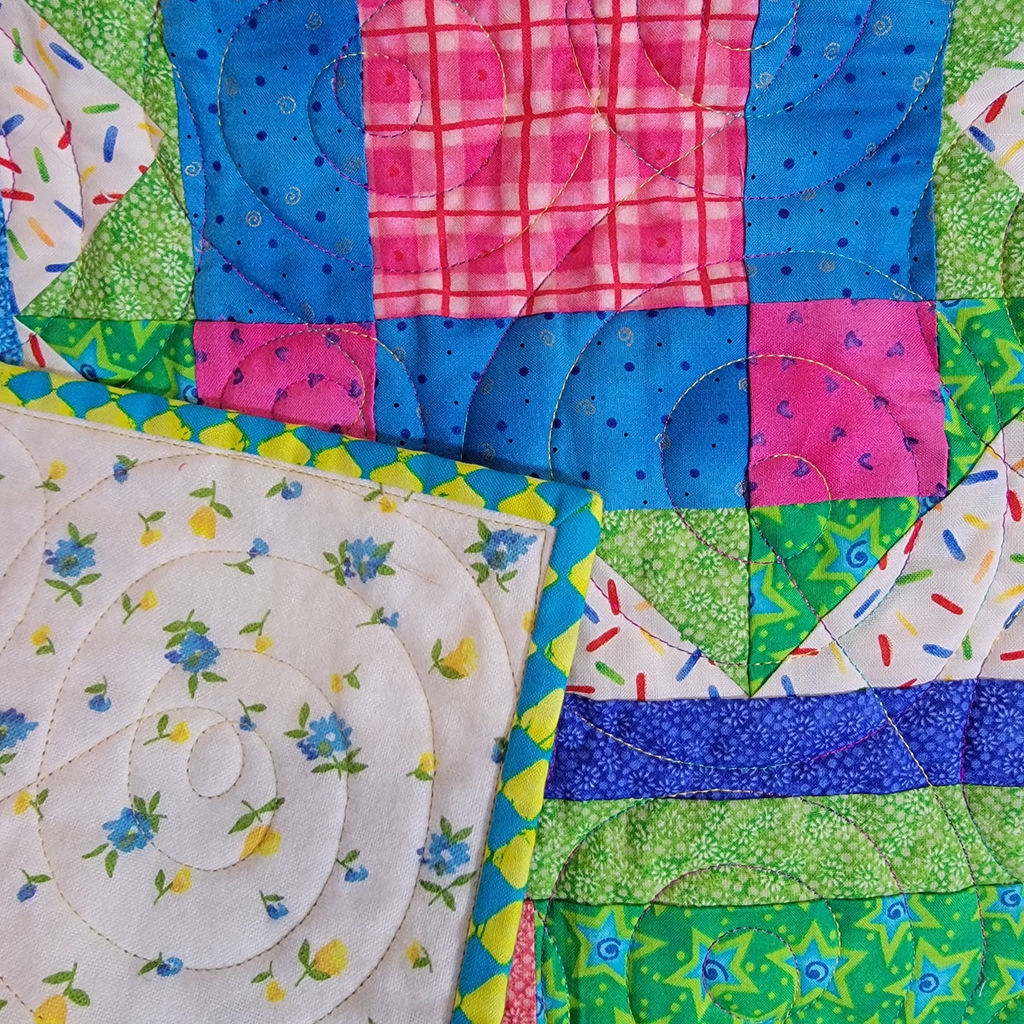 A close-up of a colorful quilted fabric with patchwork blocks in blue, pink, green, and white, including floral, plaid, and speckled patterns.