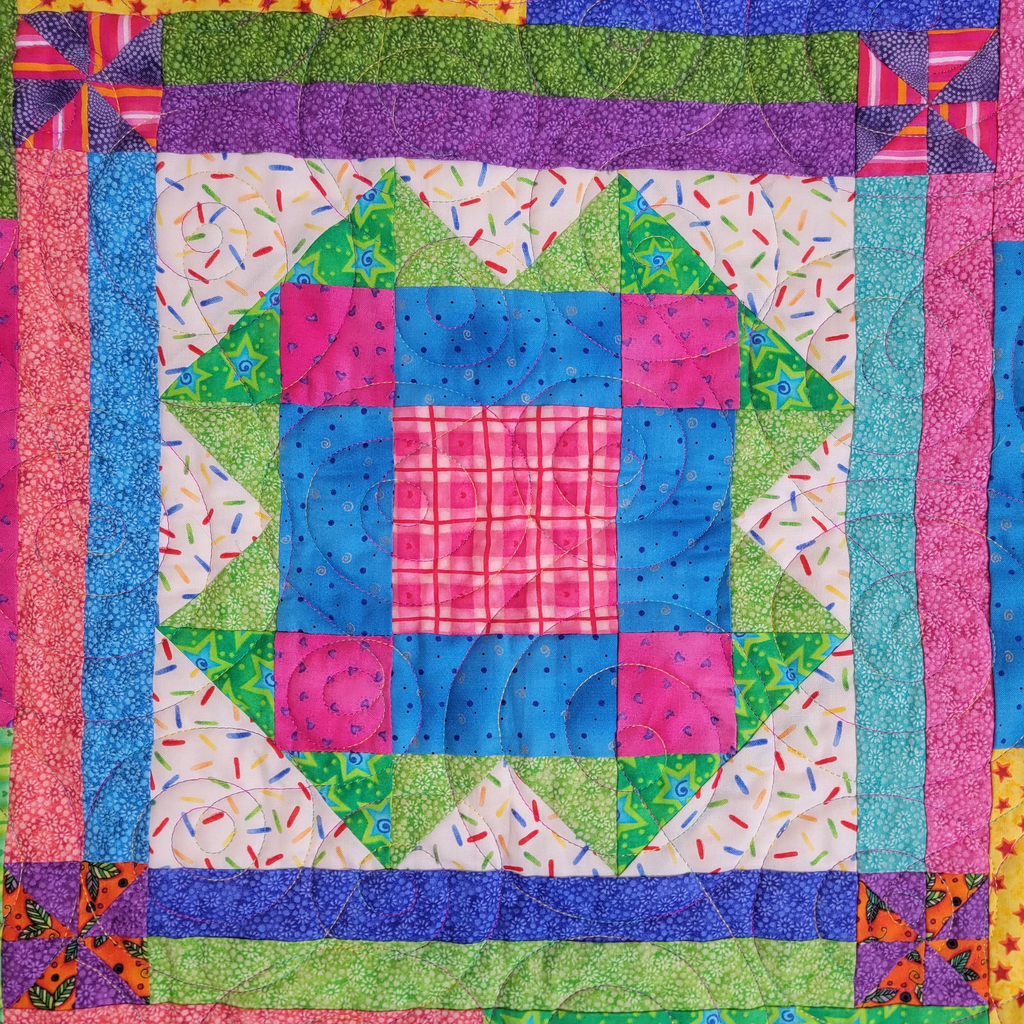 A colorful patchwork quilt featuring a central geometric block design with pink, blue, green, and white fabrics, surrounded by patterned borders.