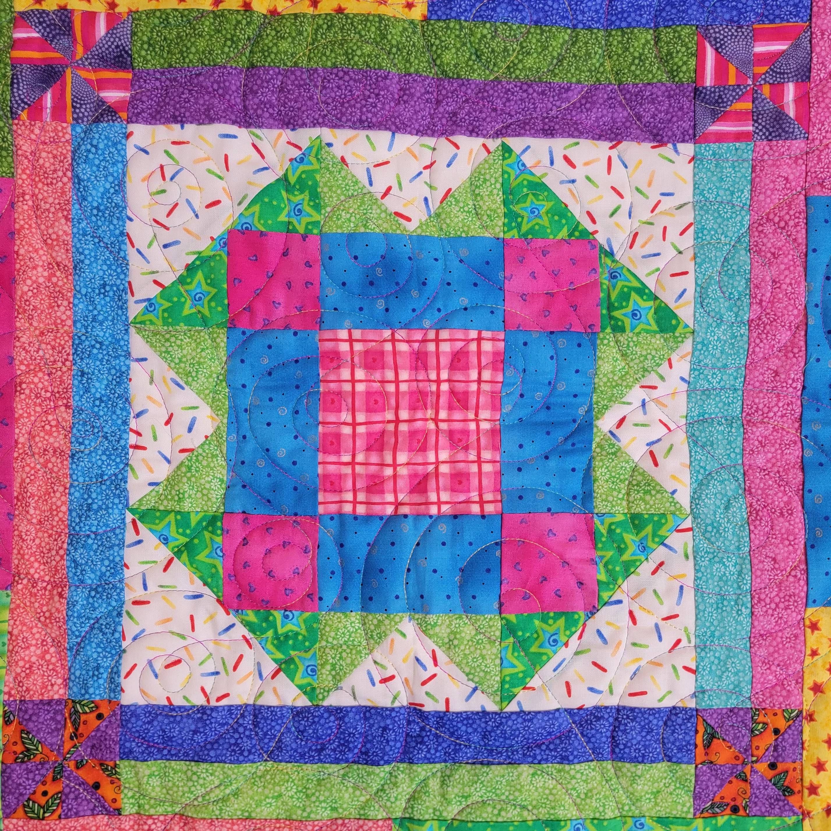 A colorful patchwork quilt featuring a central geometric block design with pink, blue, green, and white fabrics, surrounded by patterned borders.