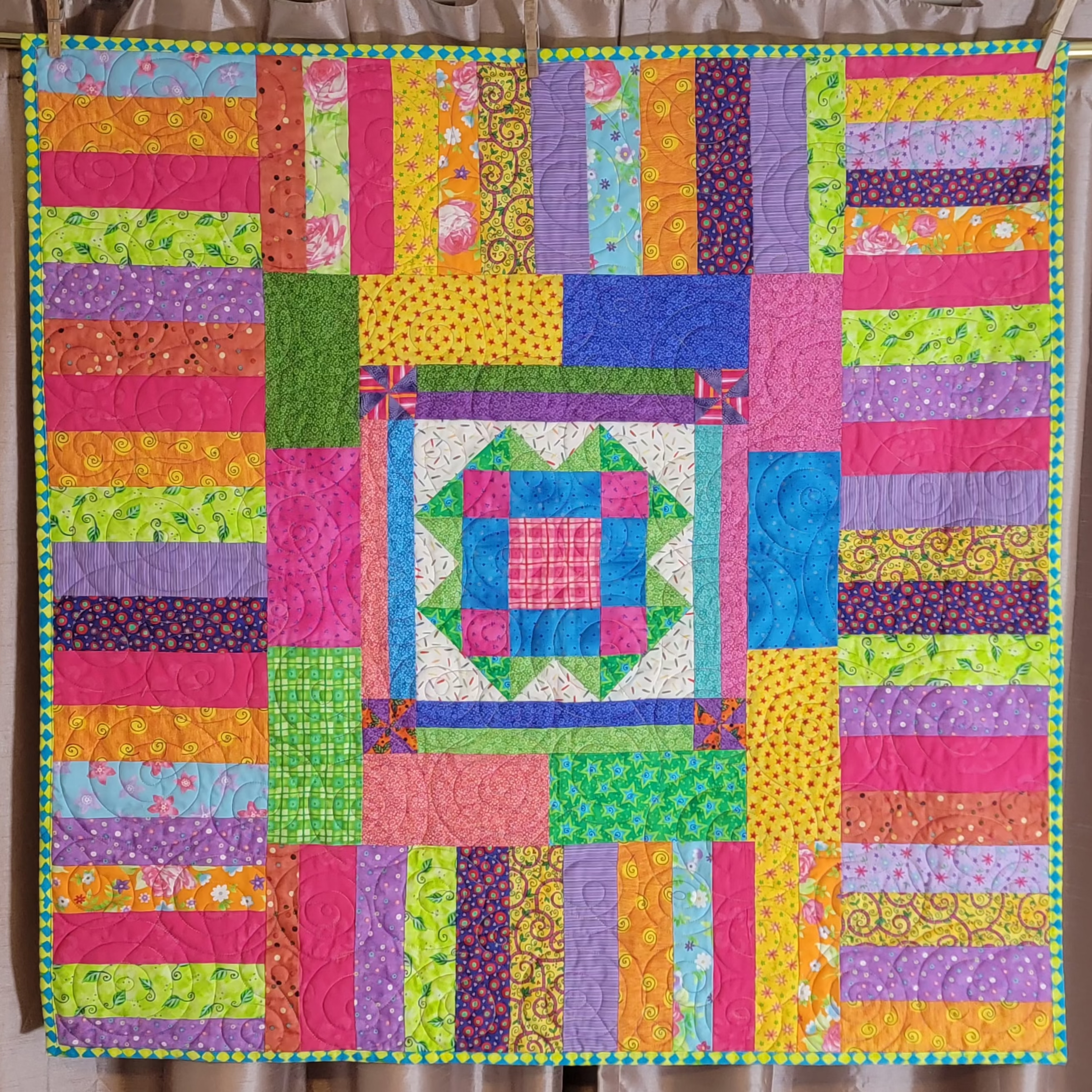 A colorful patchwork quilt hanging from a wooden rod with clothespins against a brown curtain backdrop.
