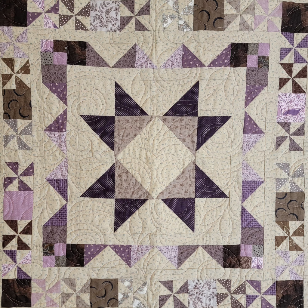 A square quilted textile featuring a central star motif in cream, beige, and purple tones, surrounded by a patterned border with purple, brown, and beige fabric sections.