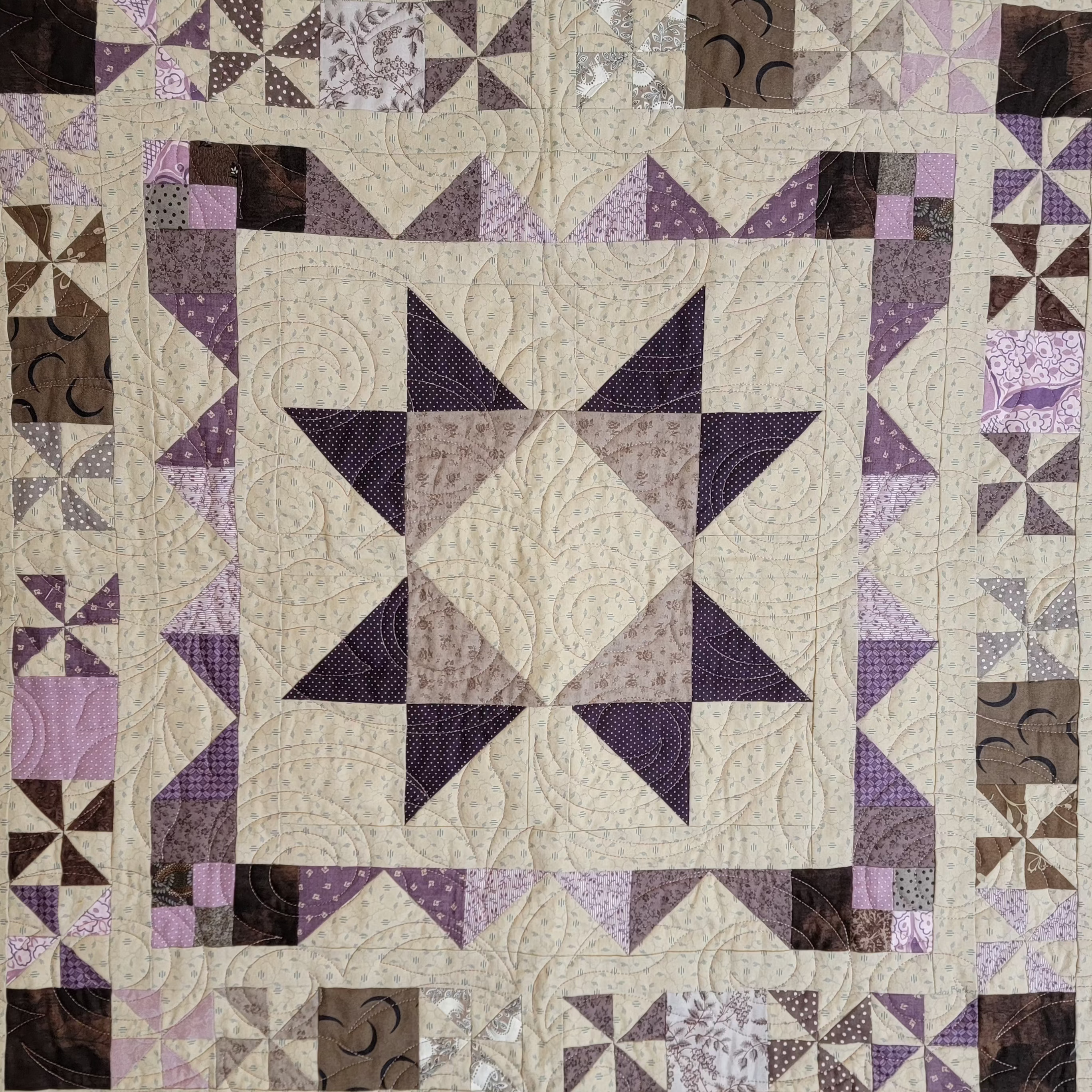A square quilted textile featuring a central star motif in cream, beige, and purple tones, surrounded by a patterned border with purple, brown, and beige fabric sections.
