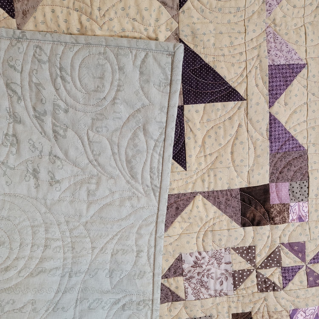 Close-up of a quilted textile with a large pale gray quilted panel on the left and a patchwork section on the right in cream, beige, brown, and purple tones with geometric and floral fabric patterns.