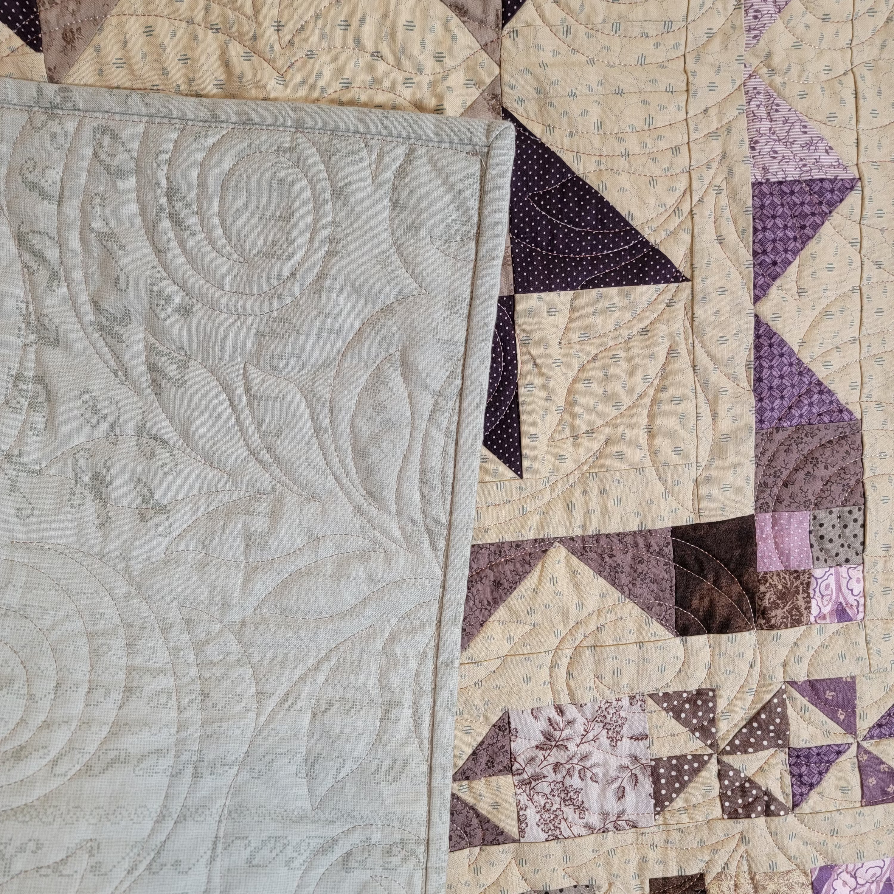 Close-up of a quilted textile with a large pale gray quilted panel on the left and a patchwork section on the right in cream, beige, brown, and purple tones with geometric and floral fabric patterns.