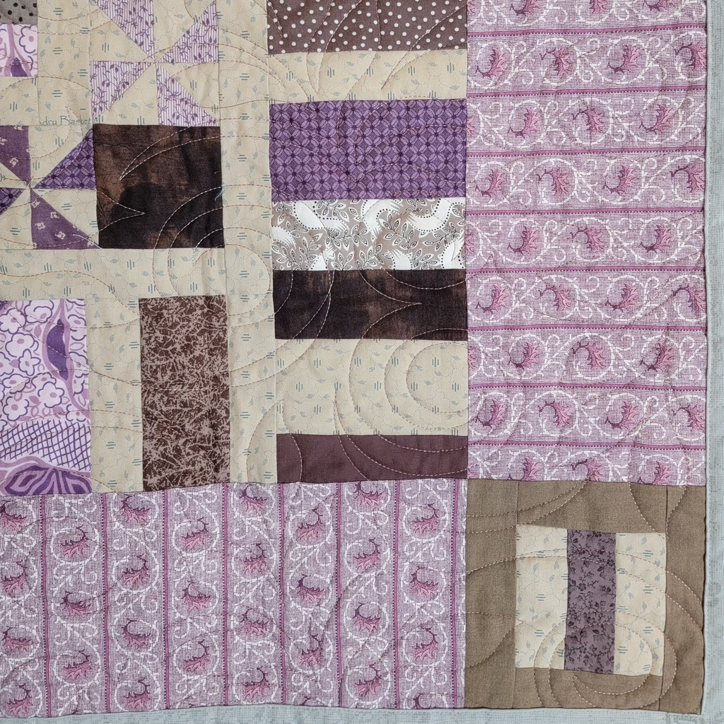 A close-up of a patchwork quilt featuring various fabric patterns in shades of purple, brown, and beige, including paisley, polka dots, and floral prints.