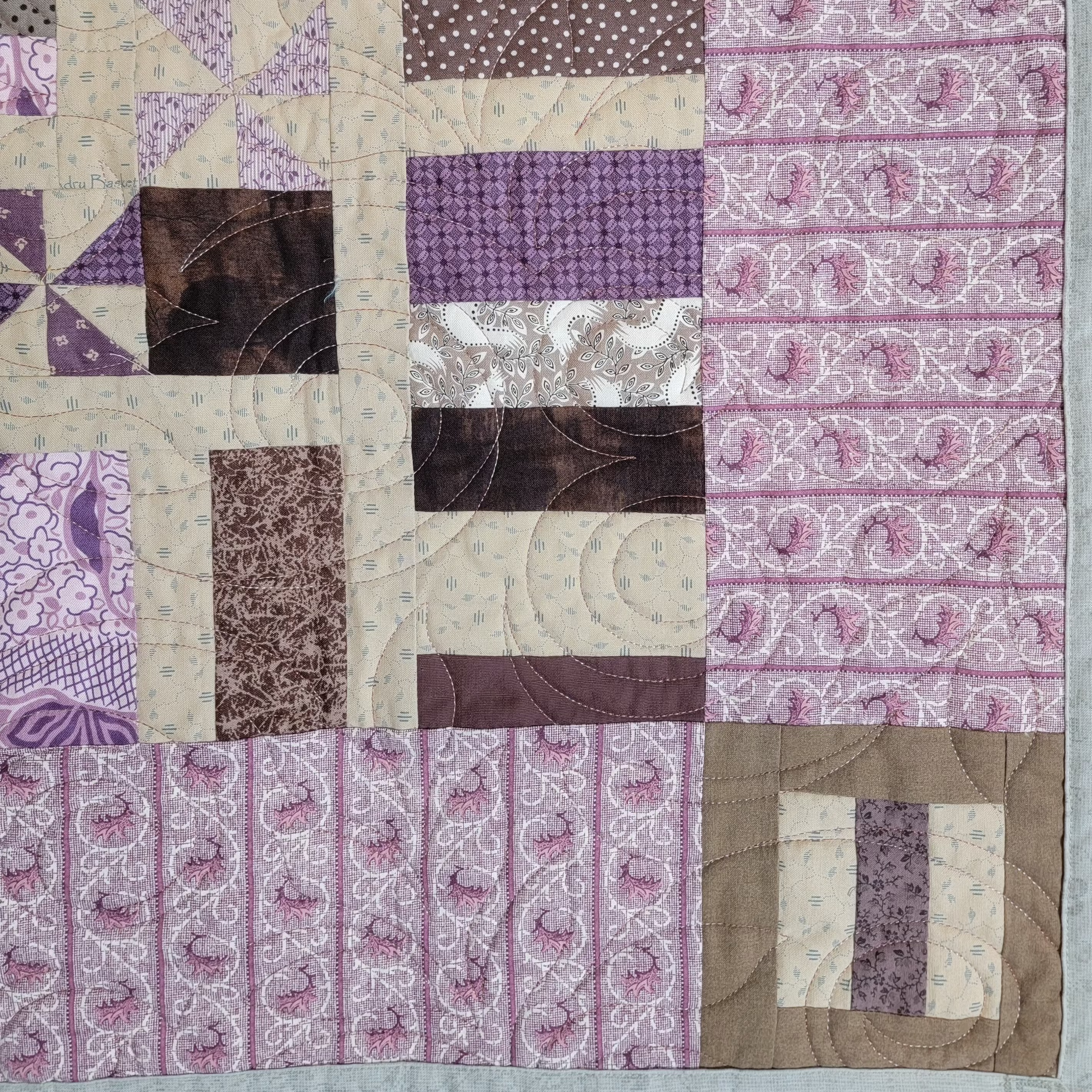 A close-up of a patchwork quilt featuring various fabric patterns in shades of purple, brown, and beige, including paisley, polka dots, and floral prints.