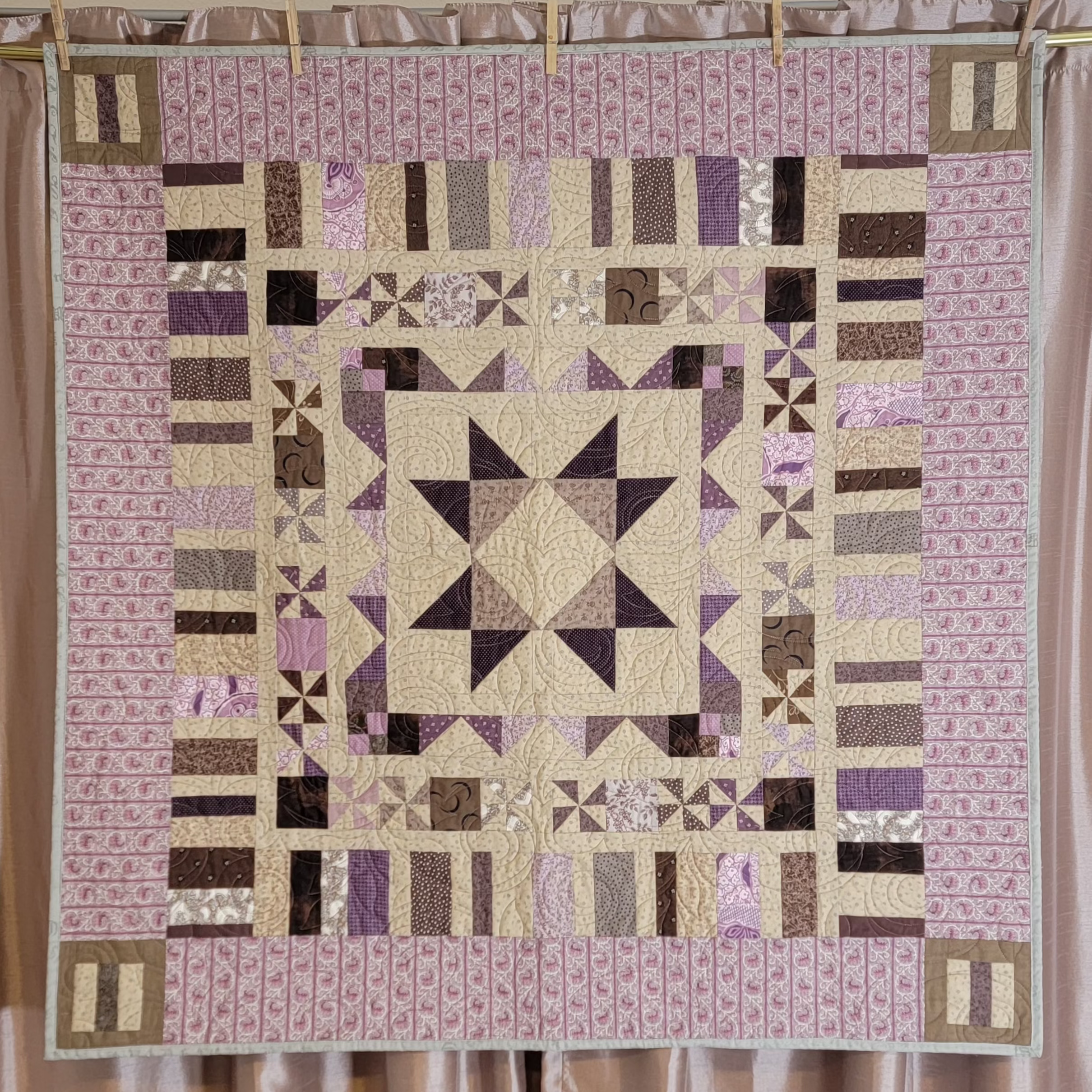 A square quilted patchwork blanket with a geometric star and striped pattern, hanging from wooden clothespins against a beige curtain backdrop.