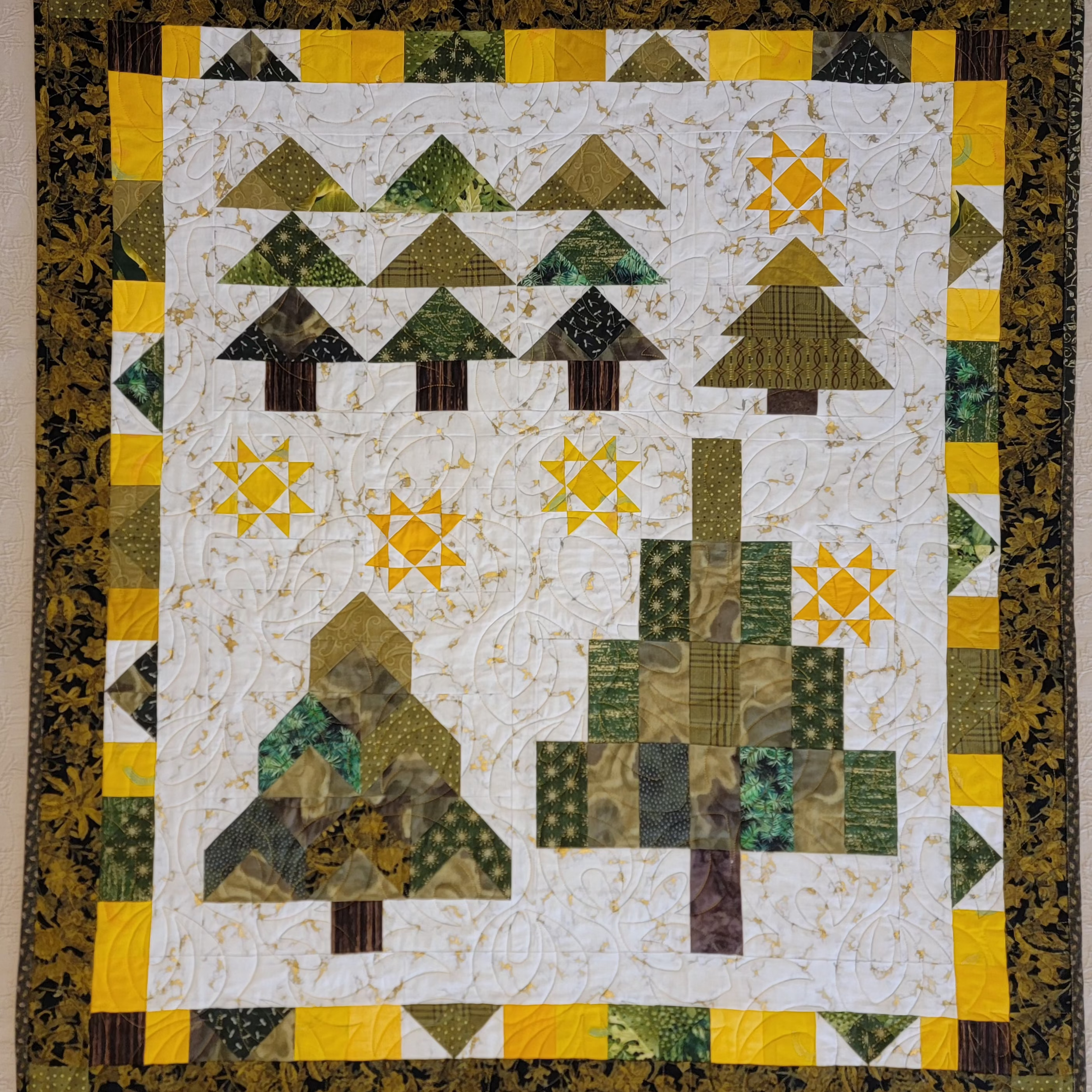 A square quilted textile featuring a stylized winter forest scene with geometric trees, starbursts, and a patterned border. The design uses olive green, mustard yellow, brown, and cream fabric pieces, photographed flat against a light neutral background.