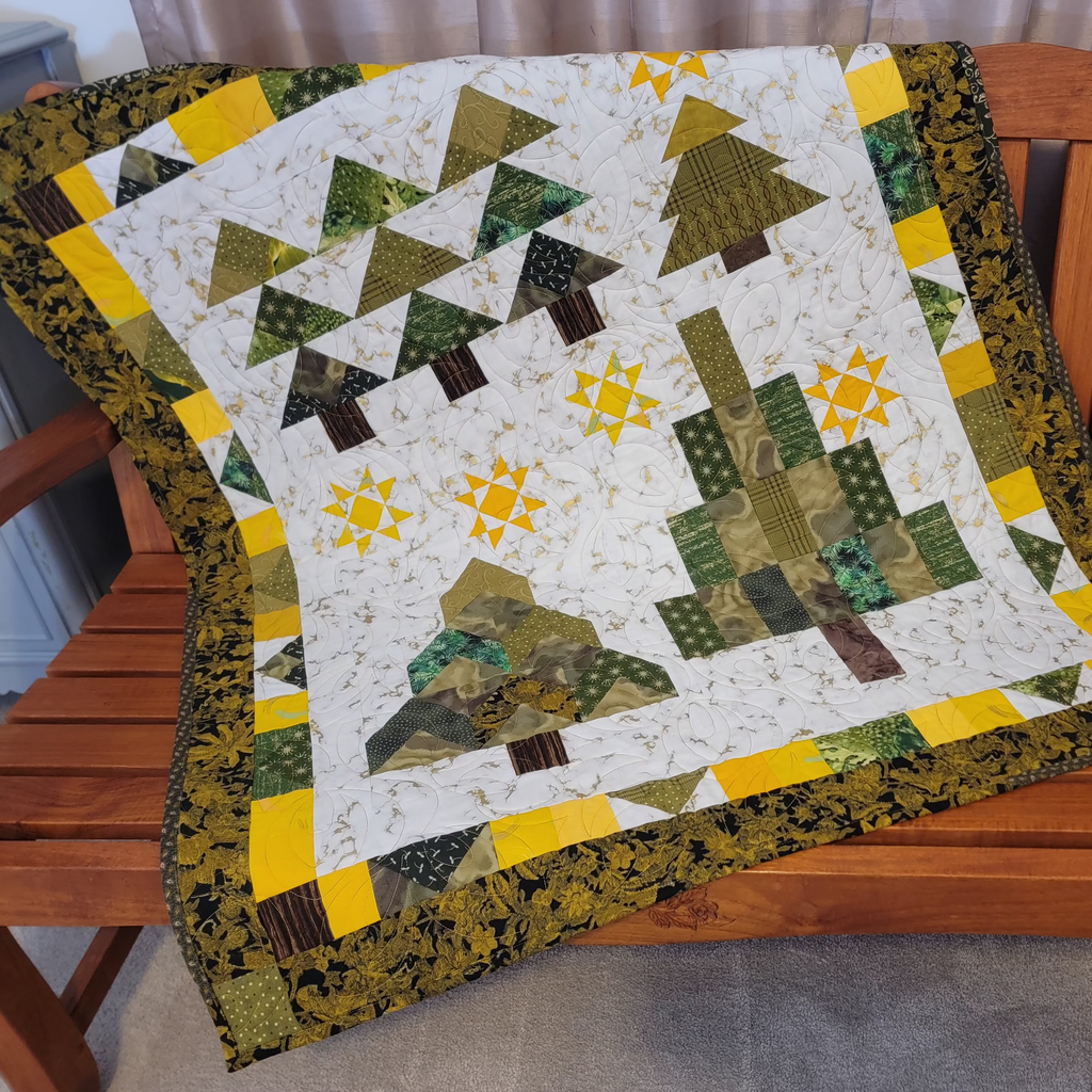 A handmade quilted patchwork blanket featuring stylized pine trees and geometric patterns in green, yellow, and brown, draped over a wooden bench.
