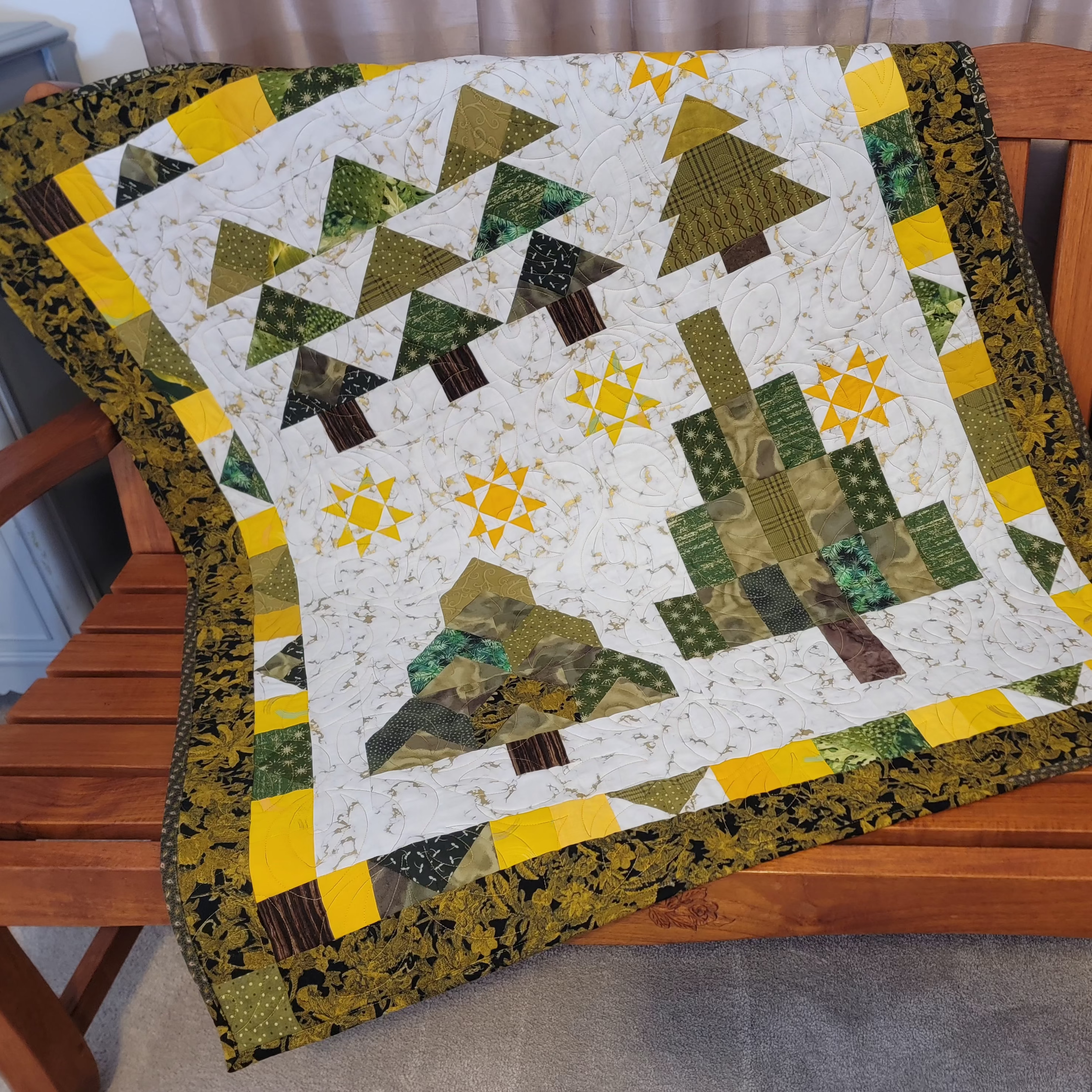 A handmade quilted patchwork blanket featuring stylized pine trees and geometric patterns in green, yellow, and brown, draped over a wooden bench.