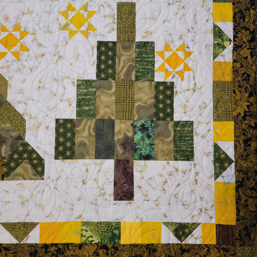 A close-up of a quilted textile featuring a tree motif made from green, olive, and brown patterned fabric blocks, surrounded by a white floral field with yellow starburst accents and a decorative border.
