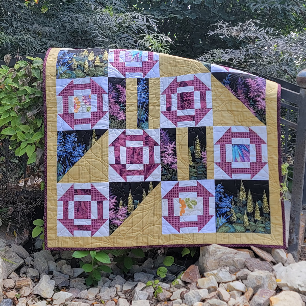 Geometric printed quilt in yellow and pinks with garden inspired prints displayed on a wooden bridge in an outdoor setting. 