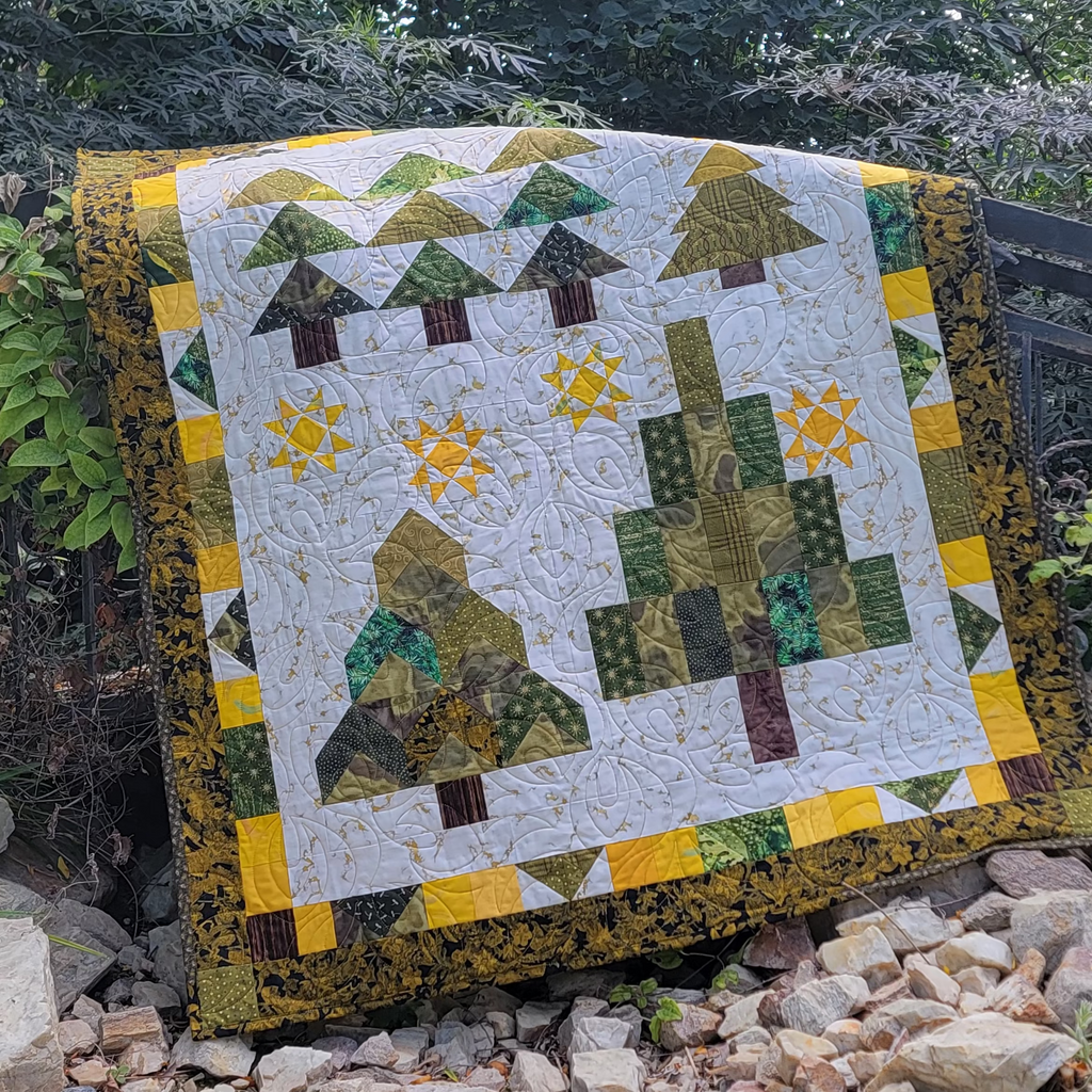 A handmade quilted patchwork blanket featuring stylized pine trees and sun motifs, draped over a black metal garden fence in a rocky, green outdoor garden setting.