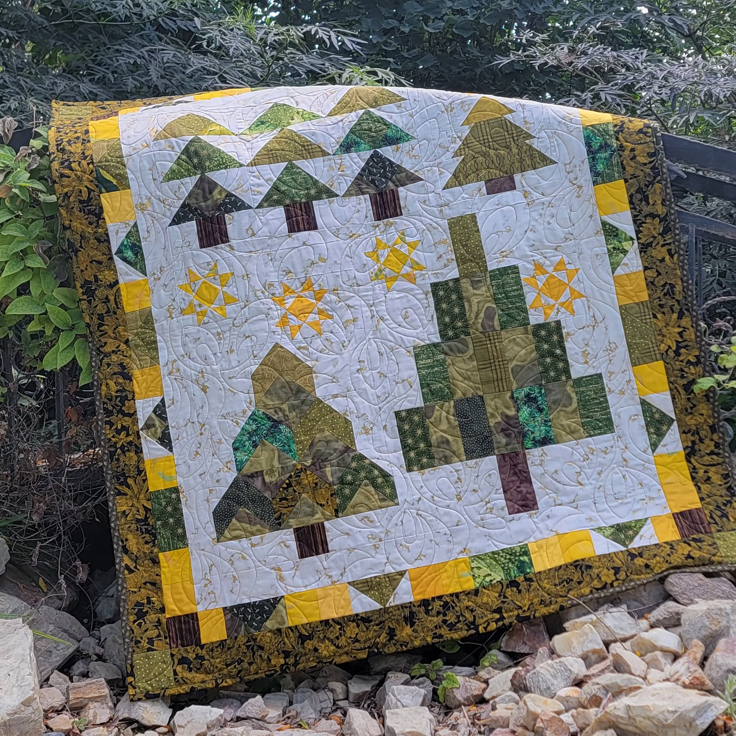 A handmade quilted patchwork blanket featuring stylized pine trees and sun motifs, draped over a black metal garden fence in a rocky, green outdoor garden setting.