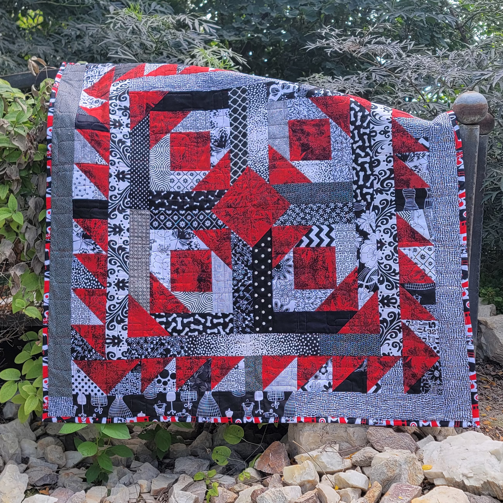A red, white and black geometric quilt draped over a wooden bridge in an outdoor setting.