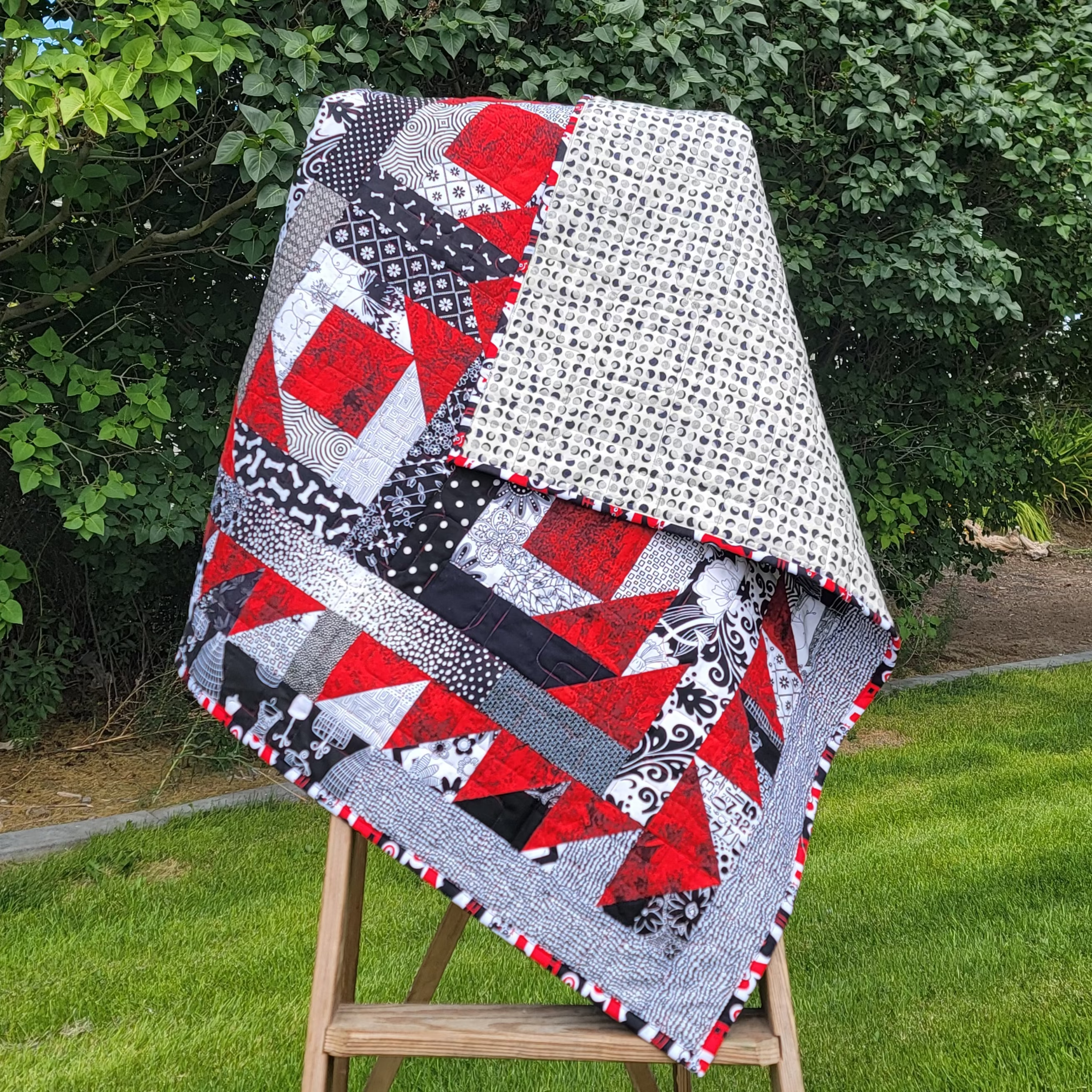 A red, white and black quilt draped over a wooden easel in an outdoor setting.