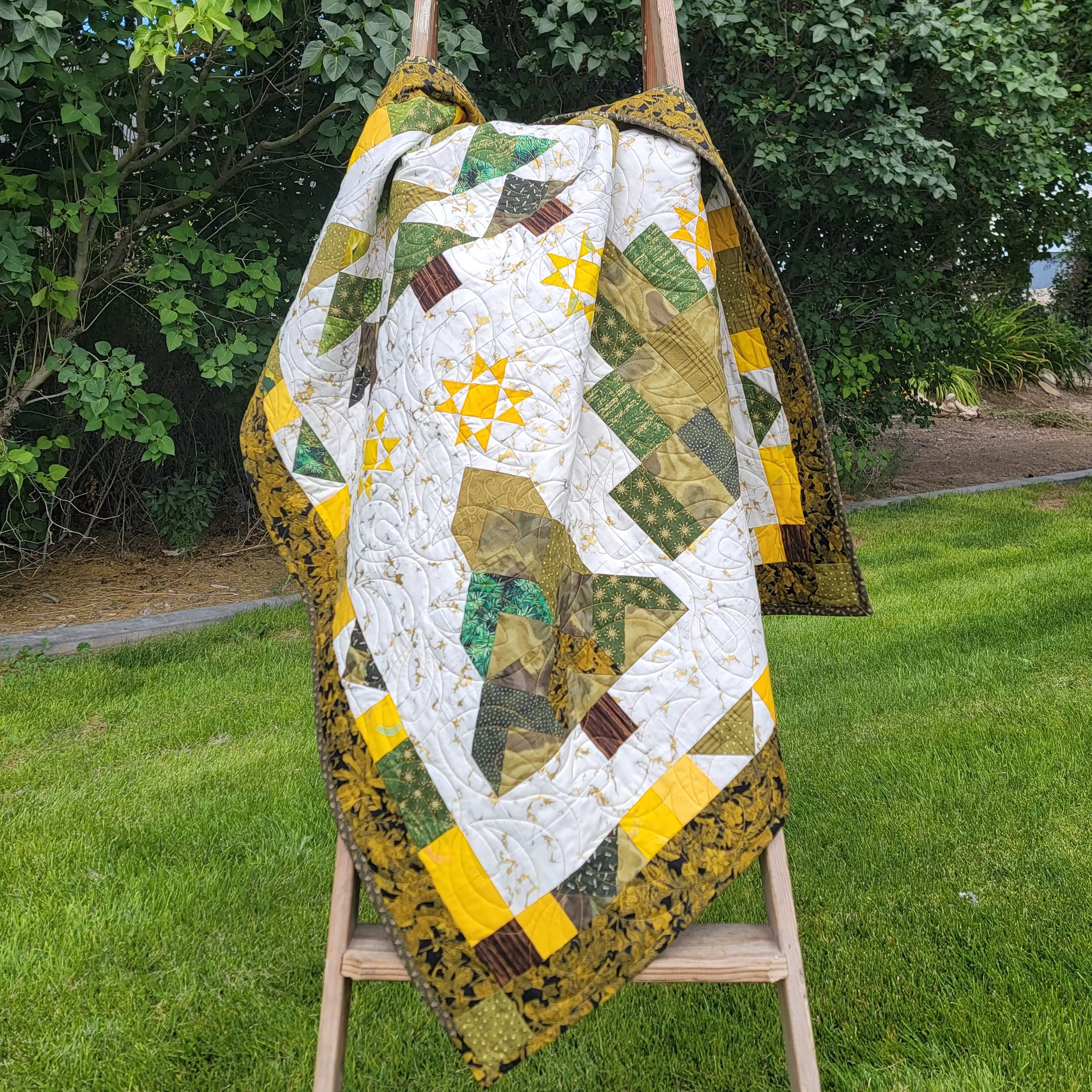 A colorful geometric quilt draped over a wooden easel in a grassy backyard with trees in the background.