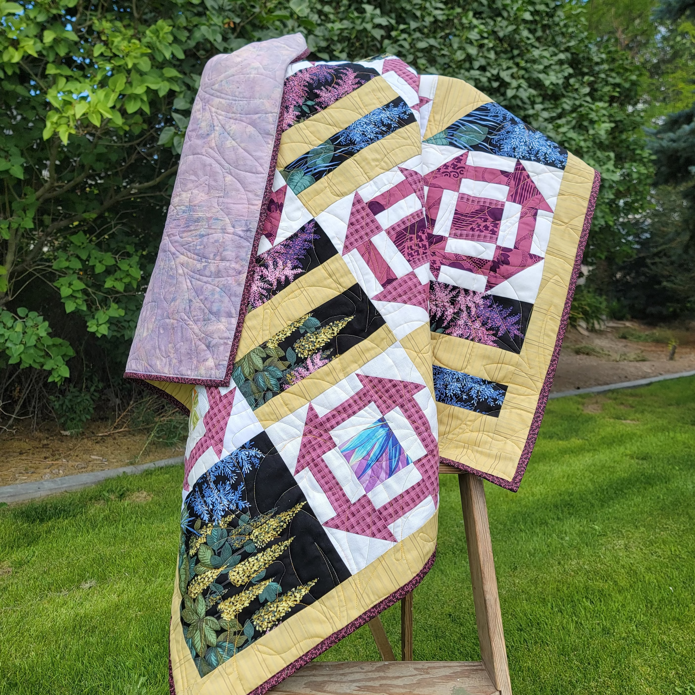 Geometric printed quilt in yellow and pinks with garden inspired prints displayed on a wooden easel in an outdoor setting. The hand dyed batik backing in purple in blue is also visible. 