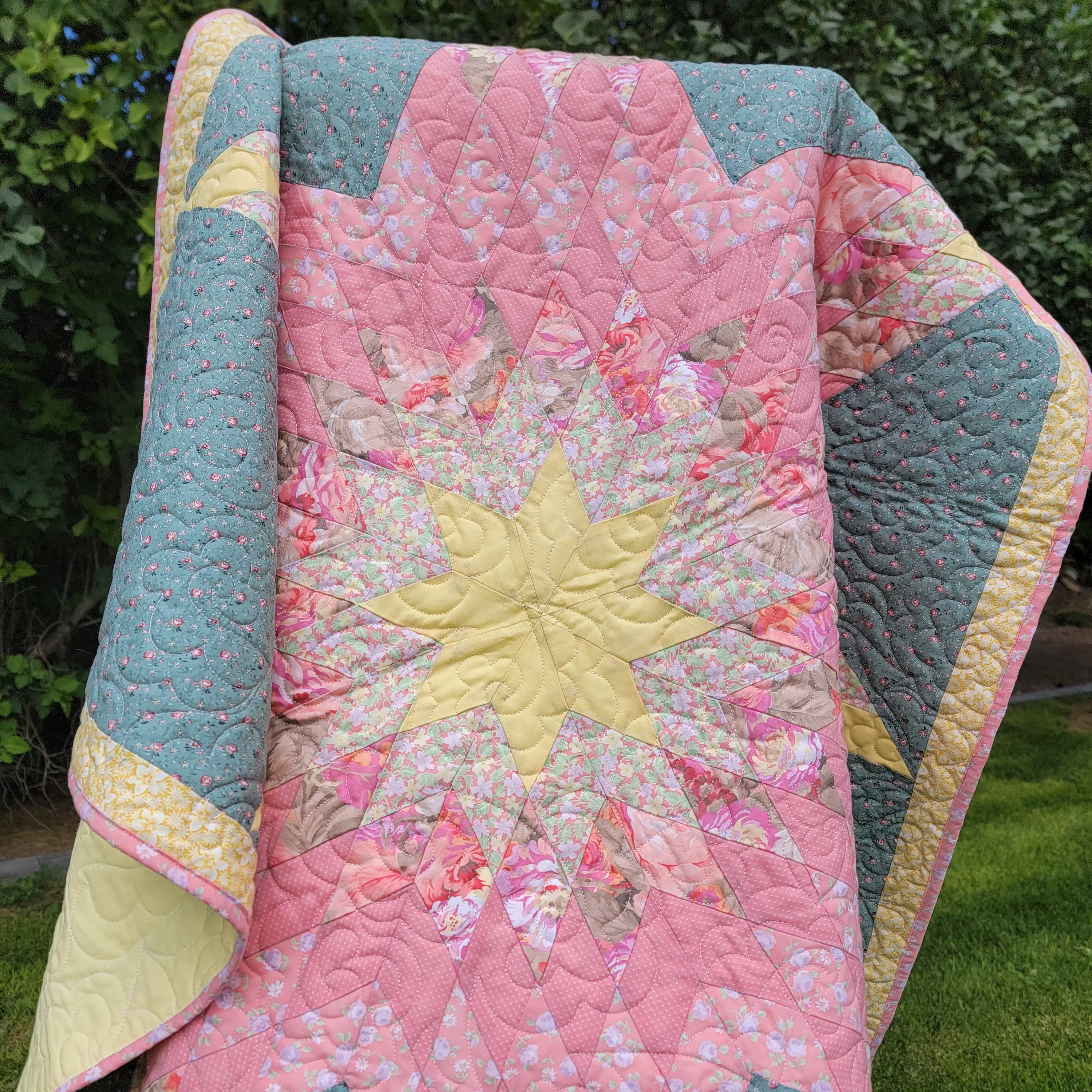 A handmade quilted starburst quilt draped outdoors on a stand, featuring a central yellow star surrounded by pink floral and patterned sections with teal and yellow borders, set against a background of green grass and foliage.