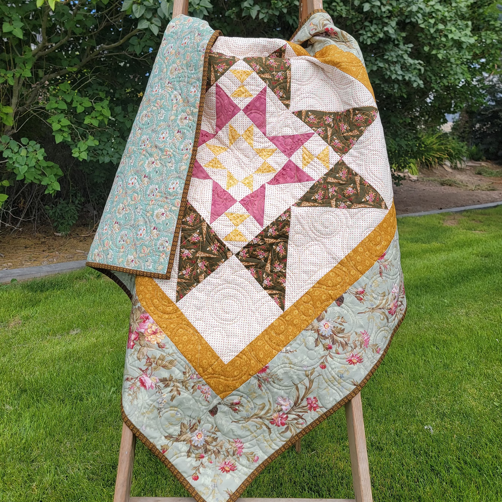 A handmade quilted star-patterned blanket draped over a wooden easel in an outdoor setting, featuring a floral border and geometric star design.