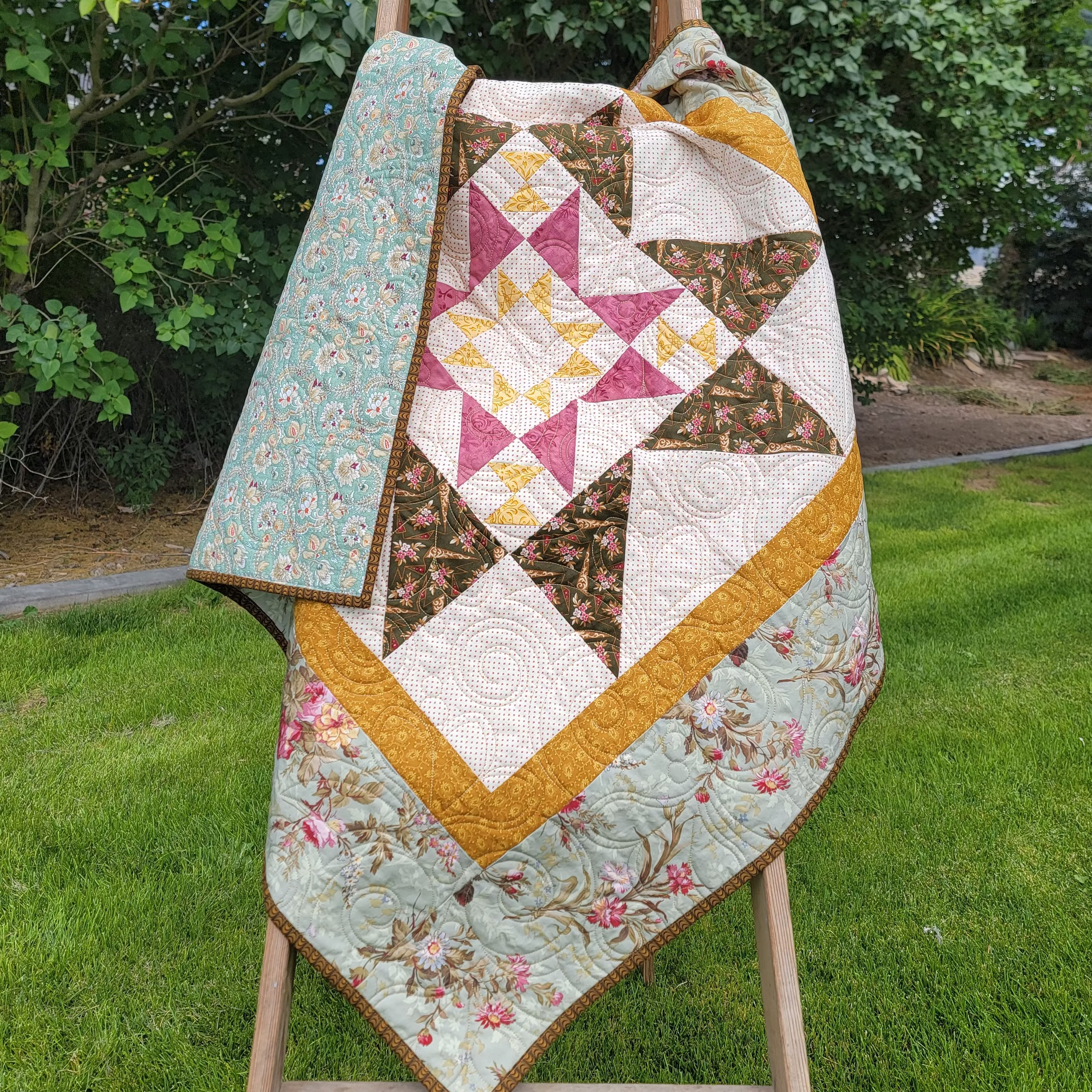 A handmade quilted star-patterned blanket draped over a wooden easel in an outdoor setting, featuring a floral border and geometric star design.