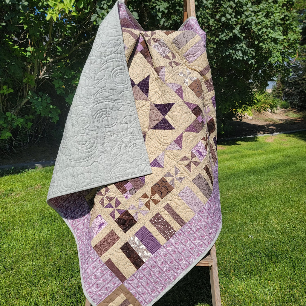 A handmade quilted blanket with a geometric patchwork design in purple, beige, and brown, draped over a wooden A-frame stand on a green grass lawn with trees in the background.