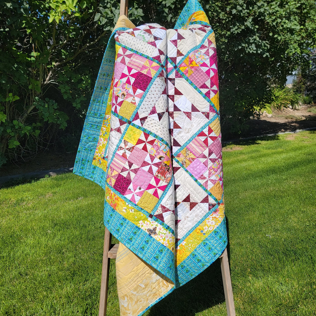A colorful patchwork quilt draped over a wooden A-frame stand in a grassy backyard with trees in the background.