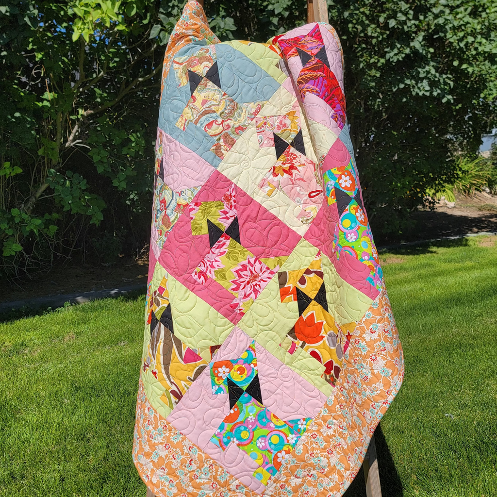 A colorful patchwork quilt with dog-themed patterns draped over a wooden easel outdoors on a green lawn with trees in the background.