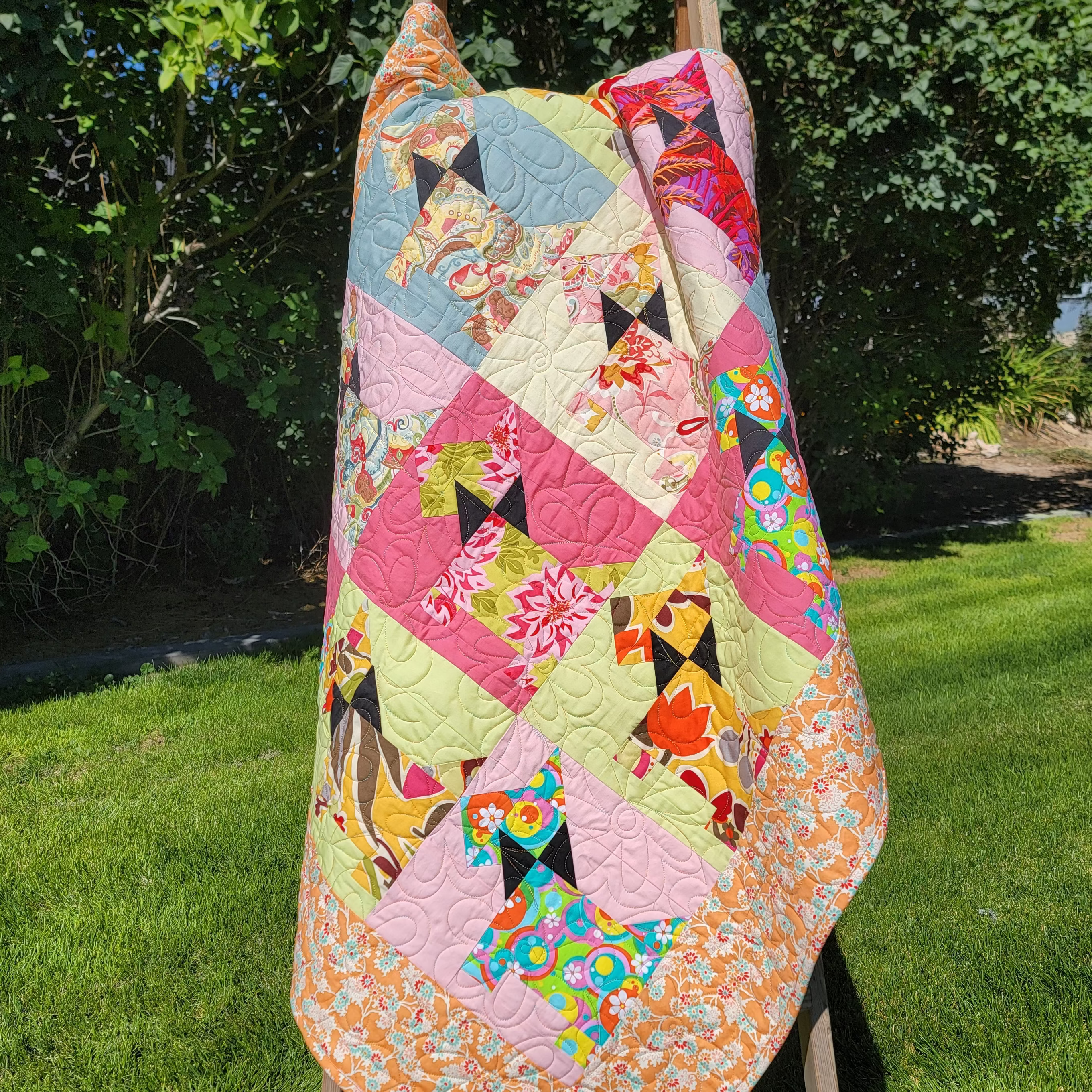 A colorful patchwork quilt with dog-themed patterns draped over a wooden easel outdoors on a green lawn with trees in the background.