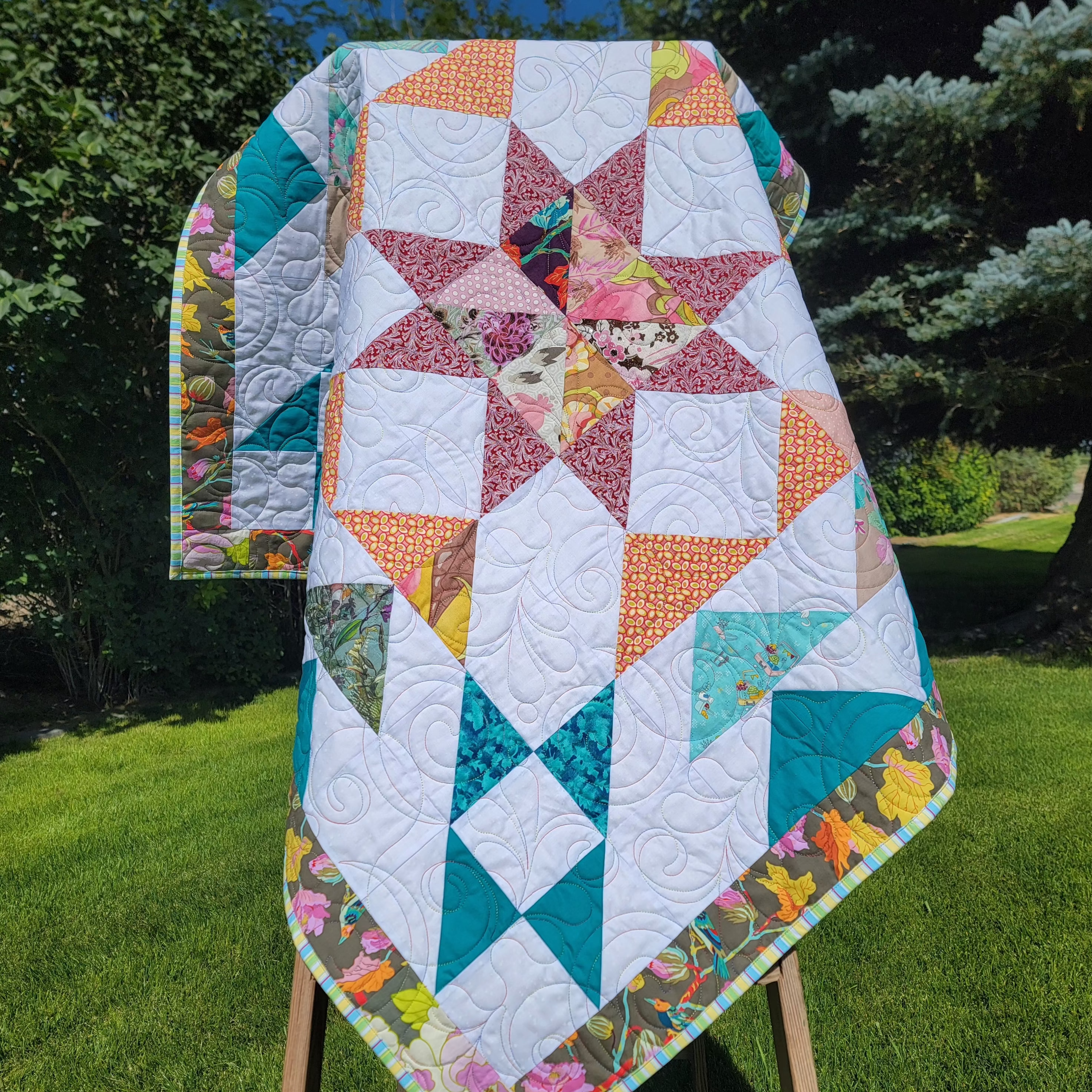 A handmade quilt in white, teal, orange and burgundy colours in multiple prints with a central star draped over a wooden easel in an outdoor setting. 
