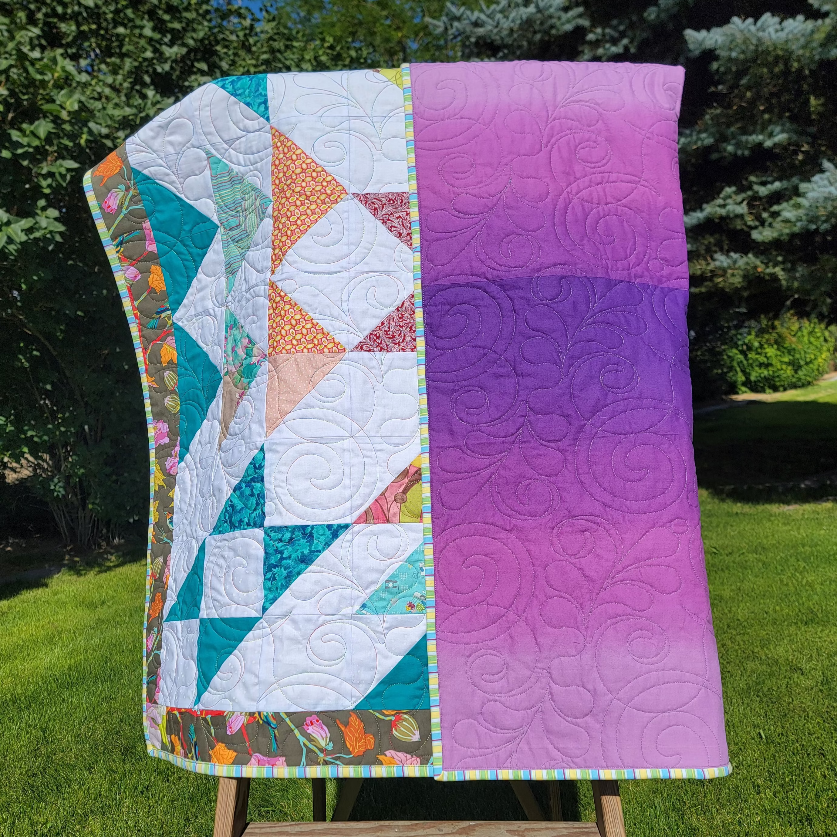 A handmade quilt in white, teal, orange and burgundy colours in multiple prints with a central star draped over a wooden easel in an outdoor setting with the reverse of the quilt showing in a purple tone with intricate stitch detail. 