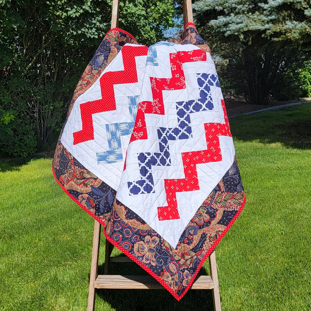 A handmade quilted baby blanket with a red, white, and blue chevron pattern and a floral border, draped over a wooden step ladder in a grassy backyard.