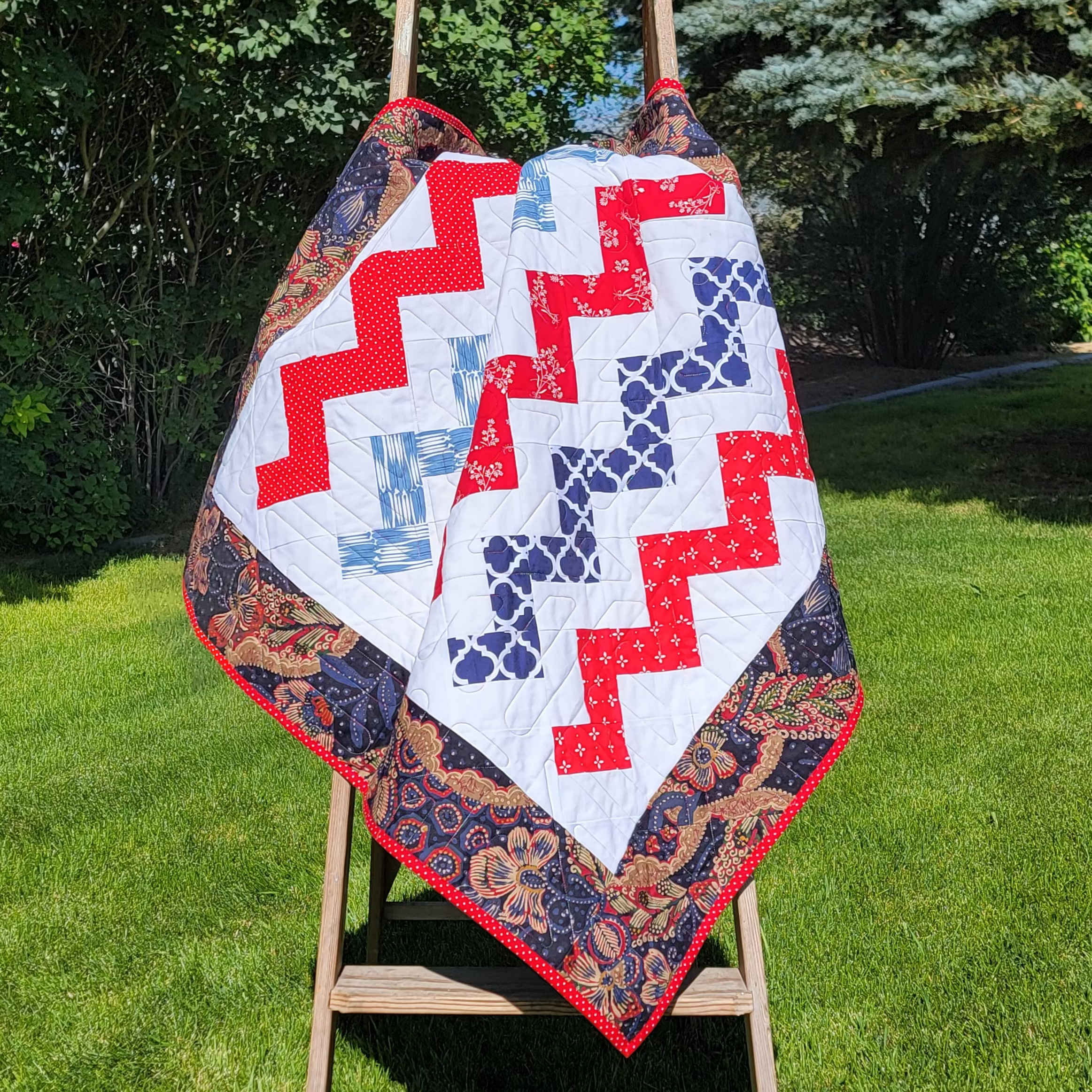 A handmade quilted baby blanket with a red, white, and blue chevron pattern and a floral border, draped over a wooden step ladder in a grassy backyard.