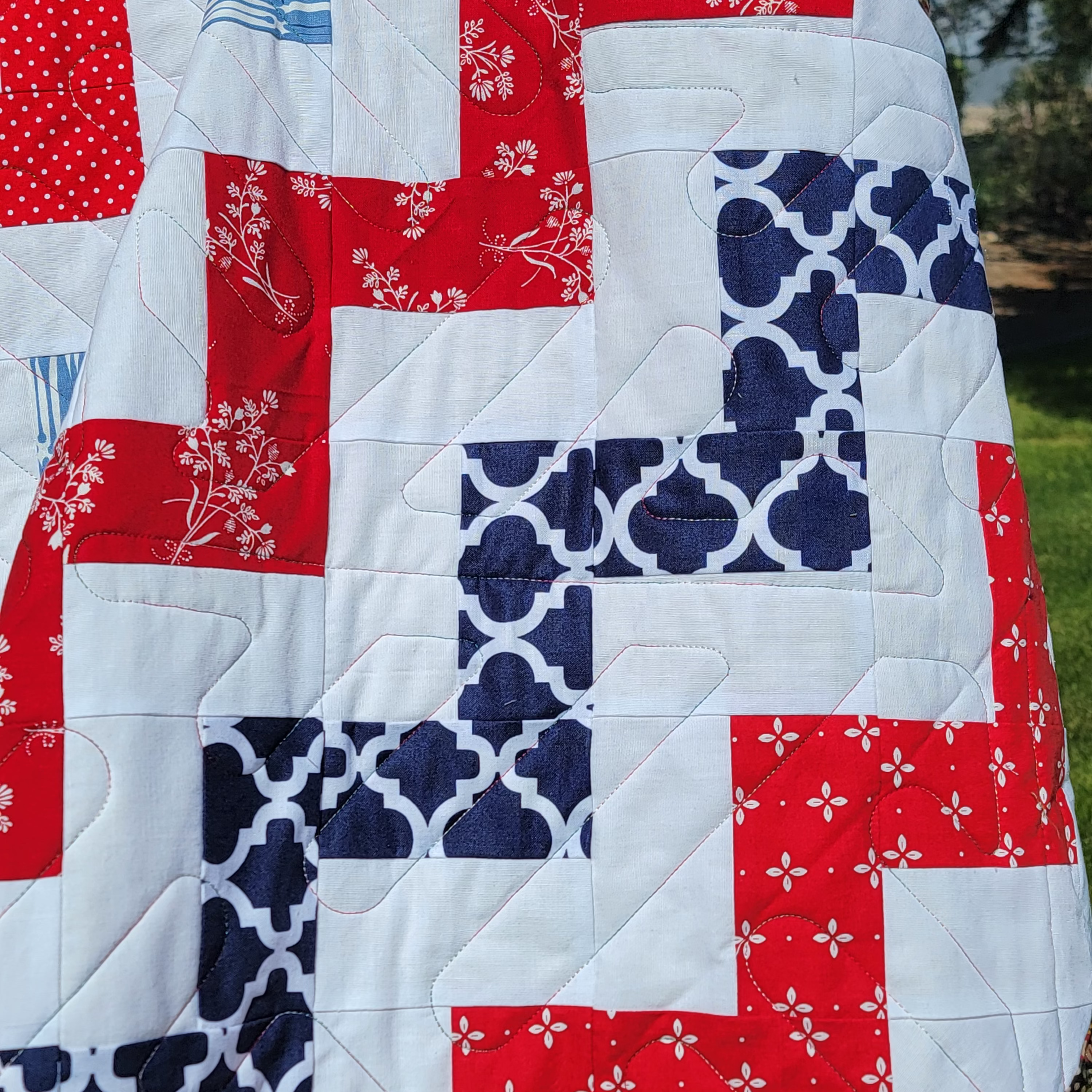 A close-up of a quilted patchwork blanket featuring red, white, and navy blue fabrics with floral and geometric patterns, draped outdoors.
